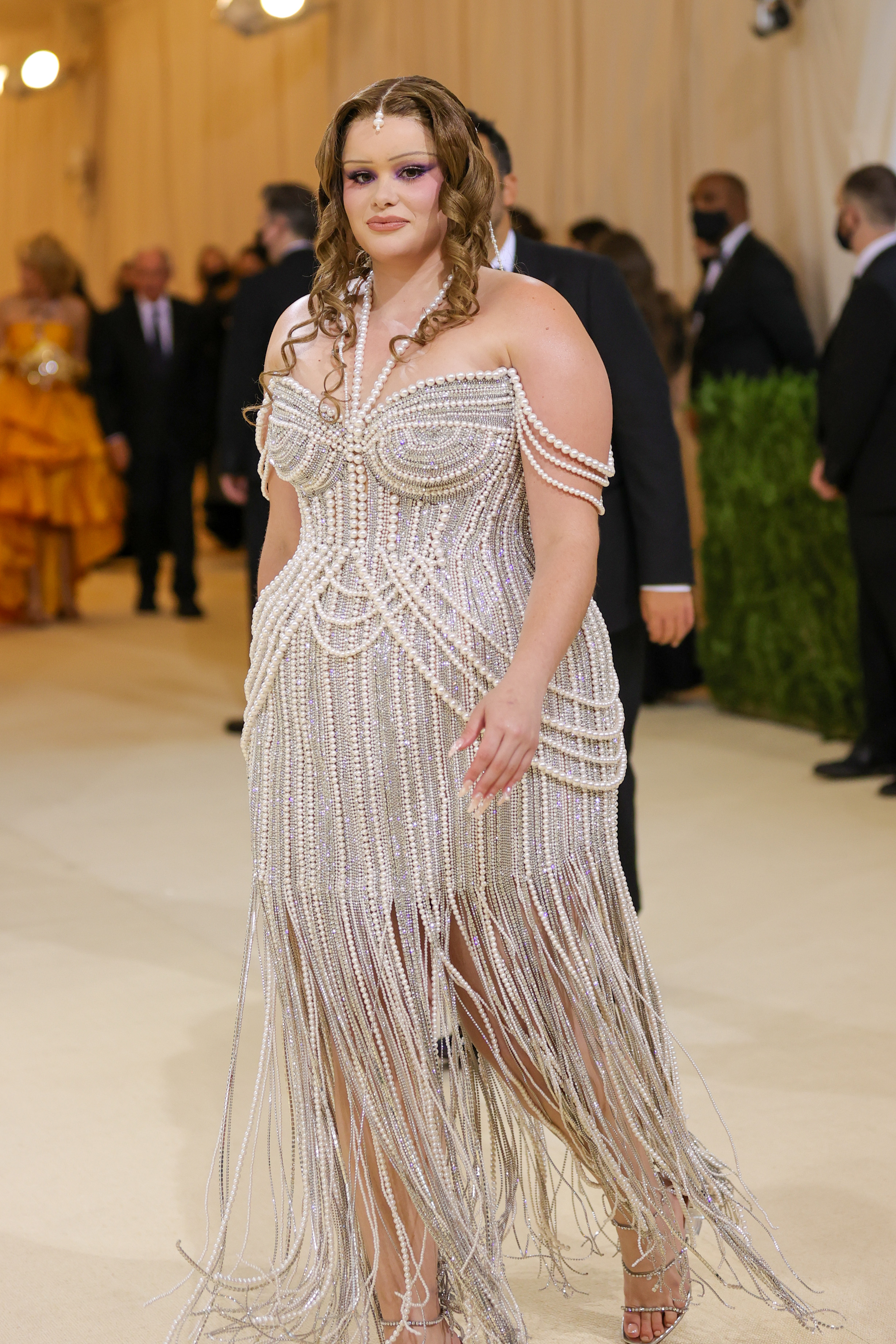 Ferreira is seen attending the 2021 Met Gala at the Metropolitan Museum of Art in New York City, celebrating “In America: A Lexicon of Fashion.” She wears an ornate pearl-embellished gown with cascading beaded fringe and off-the-shoulder draped strands. Her look is completed with glossy curls, bold lilac eye makeup, and a central forehead jewel that enhances the vintage-inspired glamour of the ensemble.