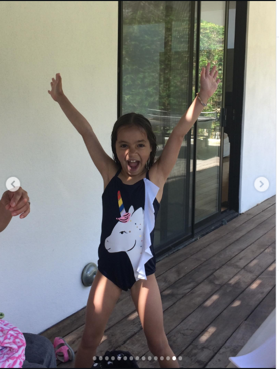 Fiona appears carefree in a playful childhood moment, standing on a wooden deck in a black one-piece swimsuit featuring a large white unicorn with a colorful horn. With both arms raised and a wide, exuberant expression, she looks full of energy as sunlight falls across the scene behind her. | Source: Instagram/mabellowe_