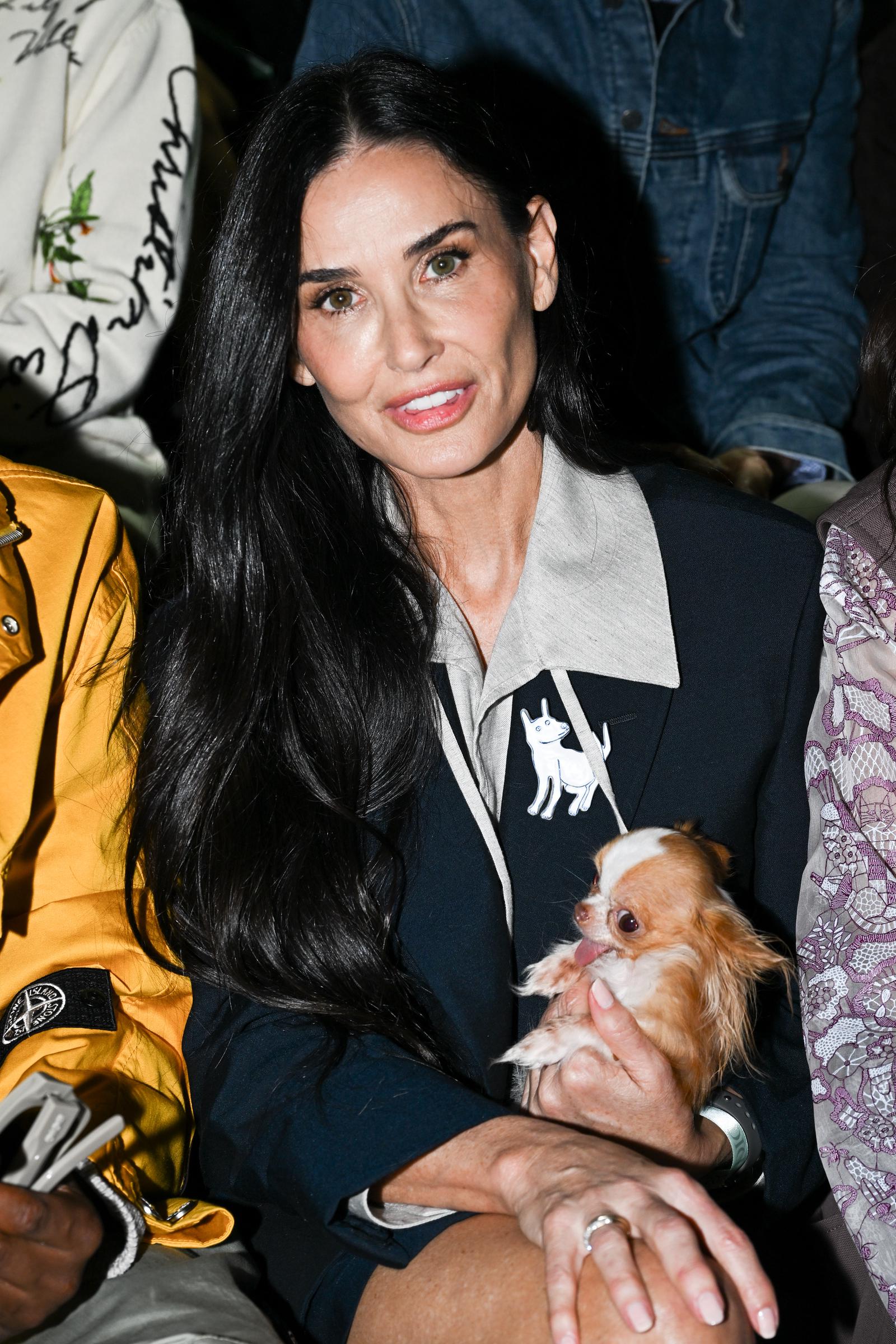 Demi Moore sits poised with a gentle smile, her hair falling softly over a chic tailored outfit accented with a playful dog motif. Cradling a tiny dog in her arms, she radiates warmth and approachability. The candid moment captures her effortless blend of elegance, charm, and genuine affection.