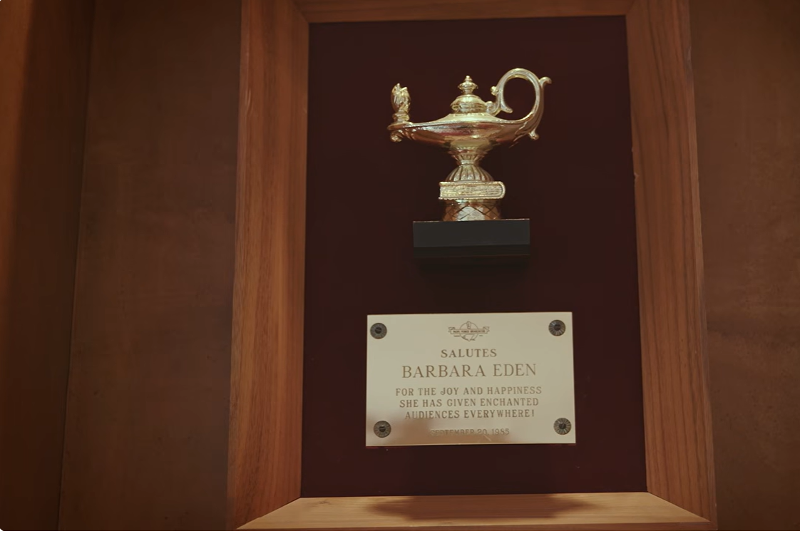 A gold-colored magical lamp sits above a commemorative plaque honoring Barbara Eden for bringing joy and happiness to audiences, dated September 20, 1985, and displayed inside a wooden cabinet. | Source: YouTube/People