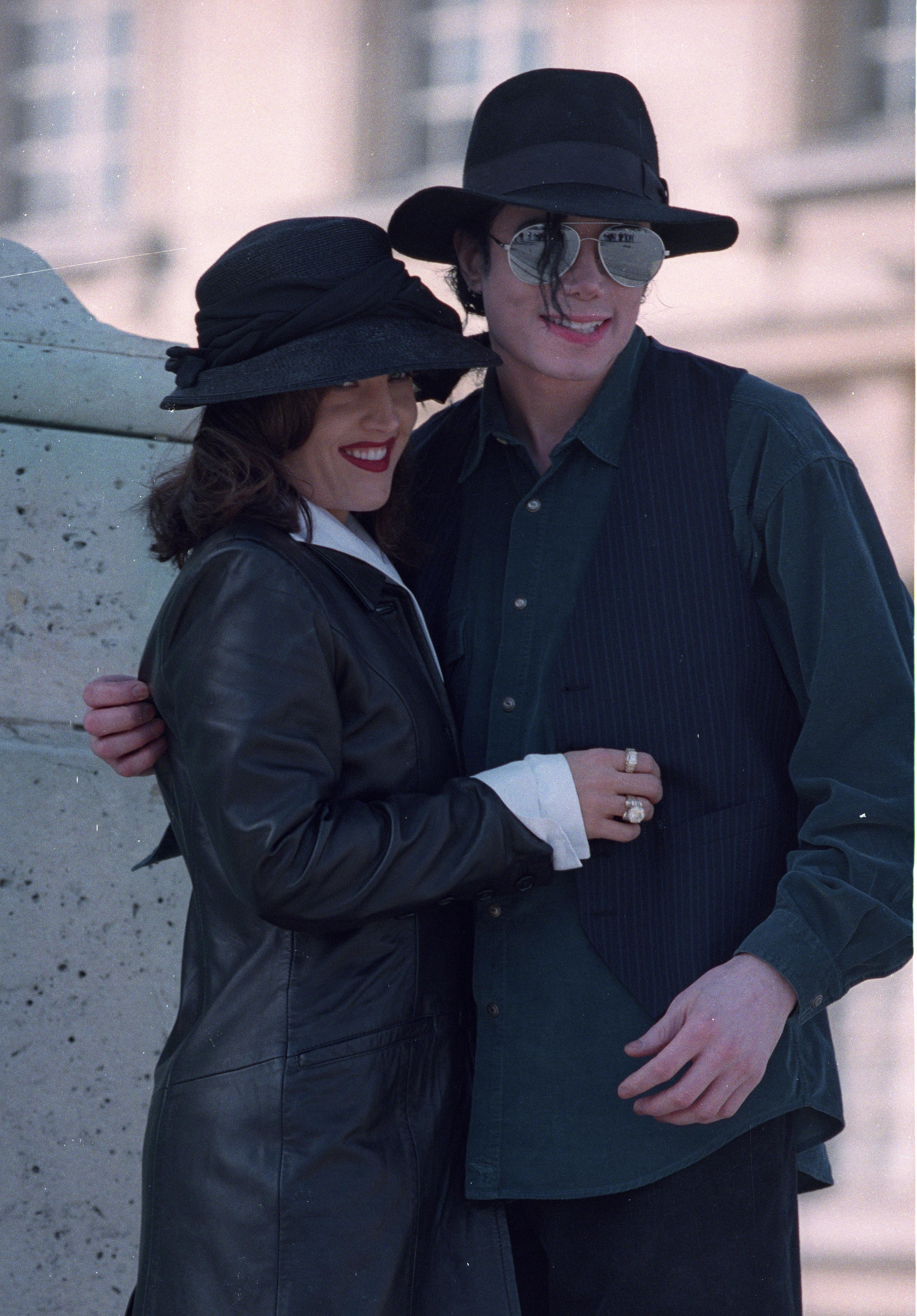 Lisa Marie Presley embraces her husband Michael Jackson during a visit to Versailles, France, in 1995, wearing a sleek black leather coat paired with a wide-brimmed black hat, while Jackson smiles in aviator sunglasses, a black fedora, and a dark button-down shirt layered beneath a pinstriped vest. | Source: Getty Images