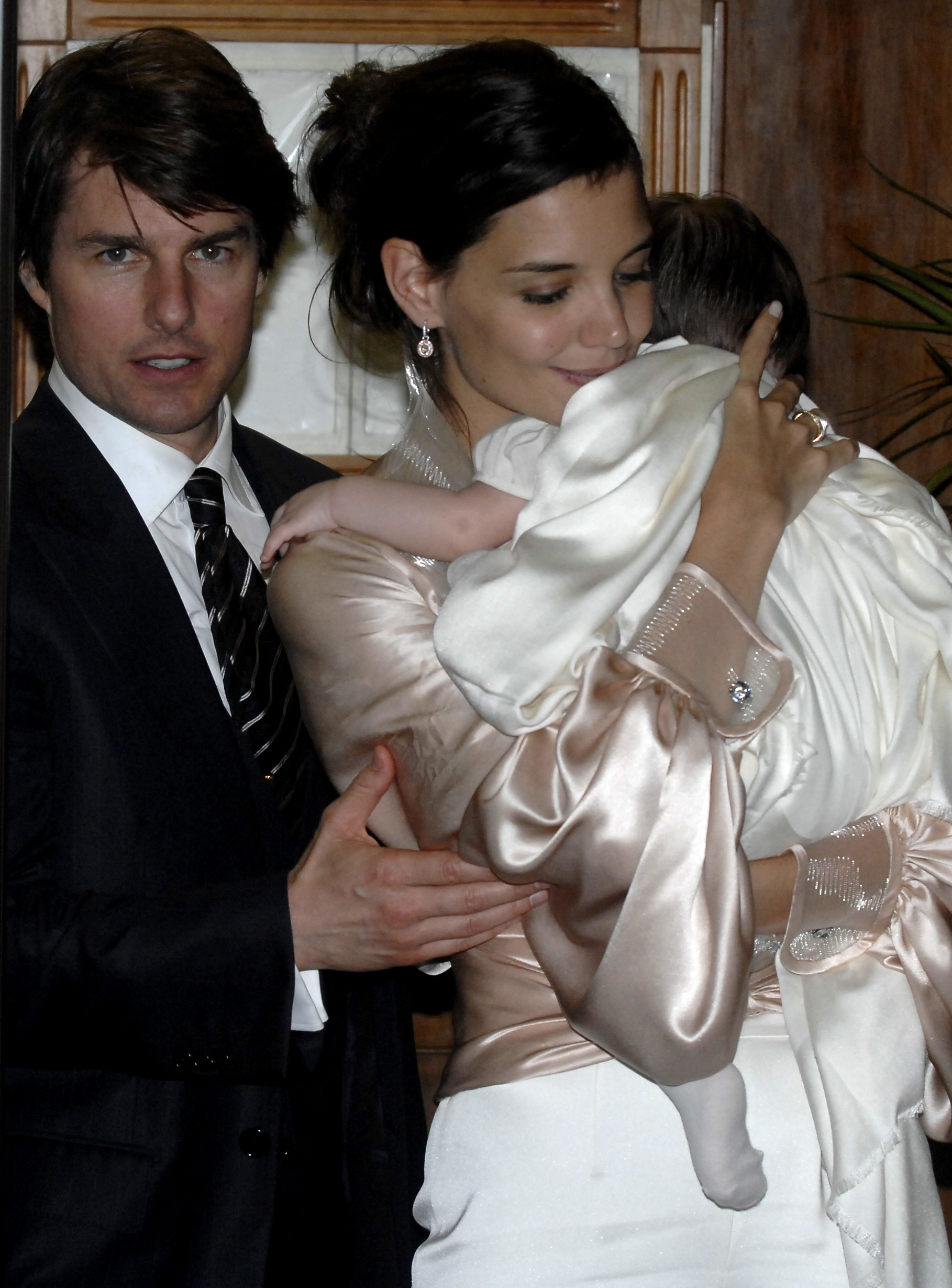 Katie Holmes lovingly cradled her baby daughter Suri, wrapped in what appears to be a satin shawl, while Tom Cruise stood protectively beside them. Dressed in elegant formal wear, the couple exuded warmth and pride.