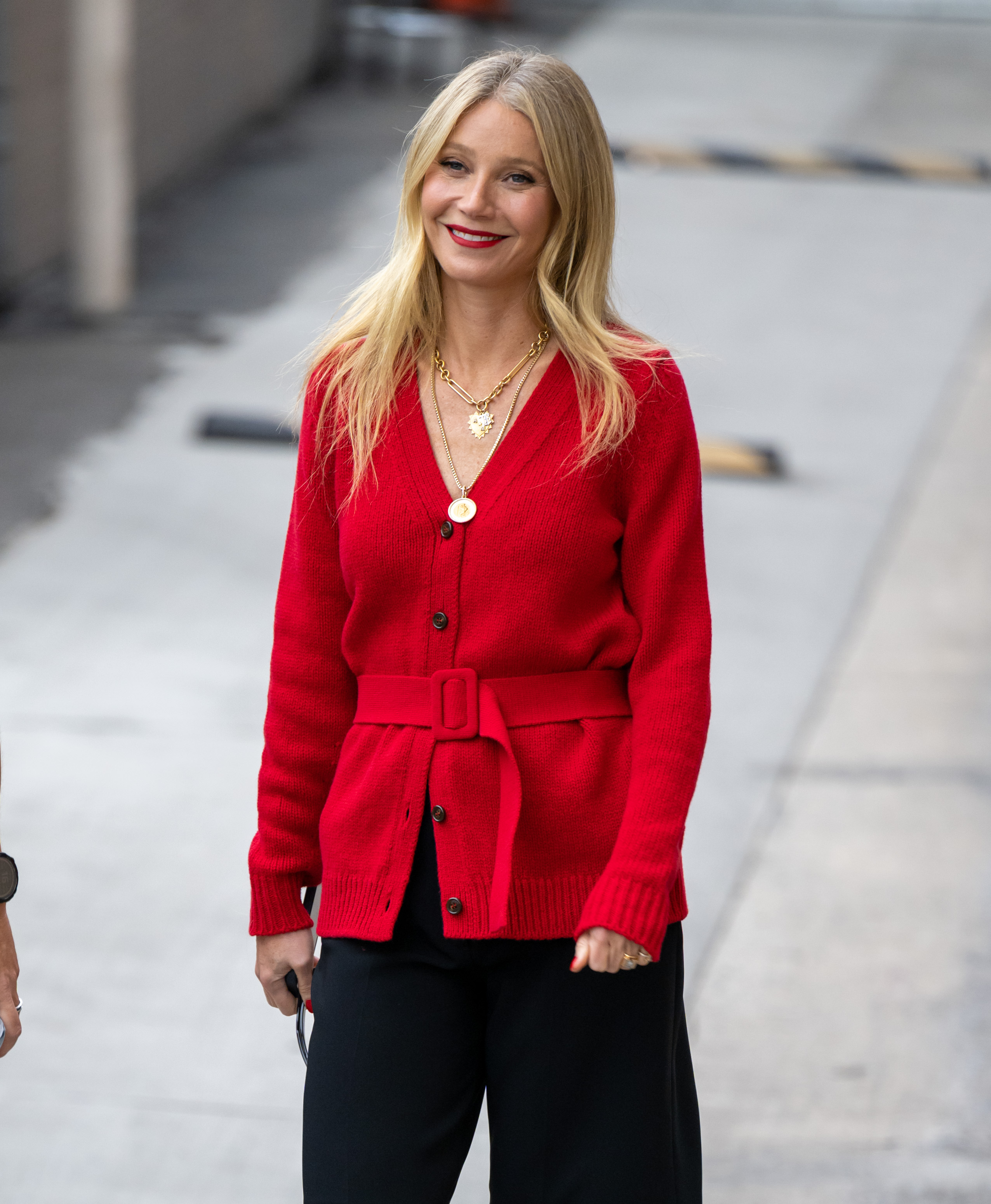 Gwyneth Paltrow arrives at "Jimmy Kimmel Live" in Los Angeles, California, on October 31, 2022. She wears a belted red cardigan layered over black trousers, paired with layered gold necklaces for a touch of shine. Her blonde hair is styled in loose waves with a center part, while a bold red lip adds a vibrant pop of color to her look.