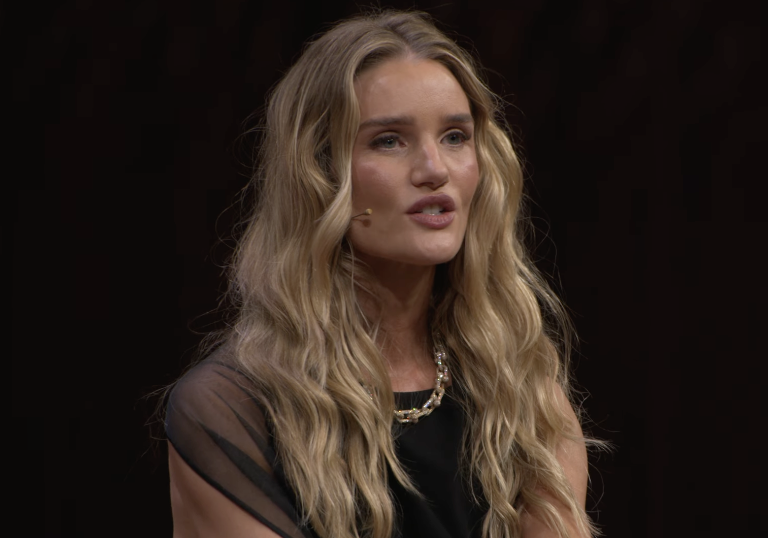 Rosie Huntington-Whiteley appears on stage mid-talk, wearing a sleek black dress with sheer detailing and a statement gold necklace. | Source: YouTube/VogueAustralia