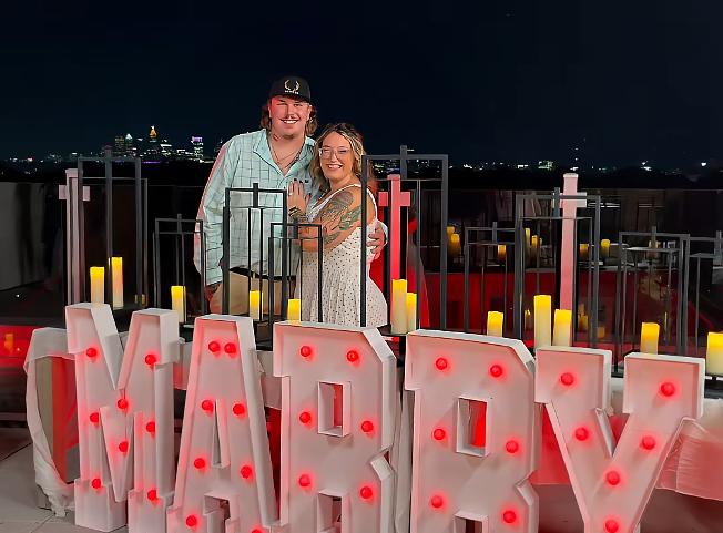 Pumpkin poses beside Darrin Kitchens in front of oversized illuminated letters spelling “MARRY,” showing off her engagement ring. The couple smiles for the camera on the rooftop where Darrin planned the romantic proposal. | Source: TikTok/Mama P
