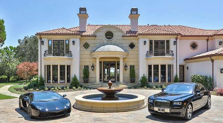 The grand cobblestone motor court of Reba McEntire's former Beverly Park mansion centers on a tiered stone fountain, flanked by luxury sports cars before the home's imposing entrance. The Italianate facade features a domed columned portico, dual chimneys, wrought-iron balustraded balconies, and classical terracotta tile rooflines. | Source: YouTube/Taste of Country
