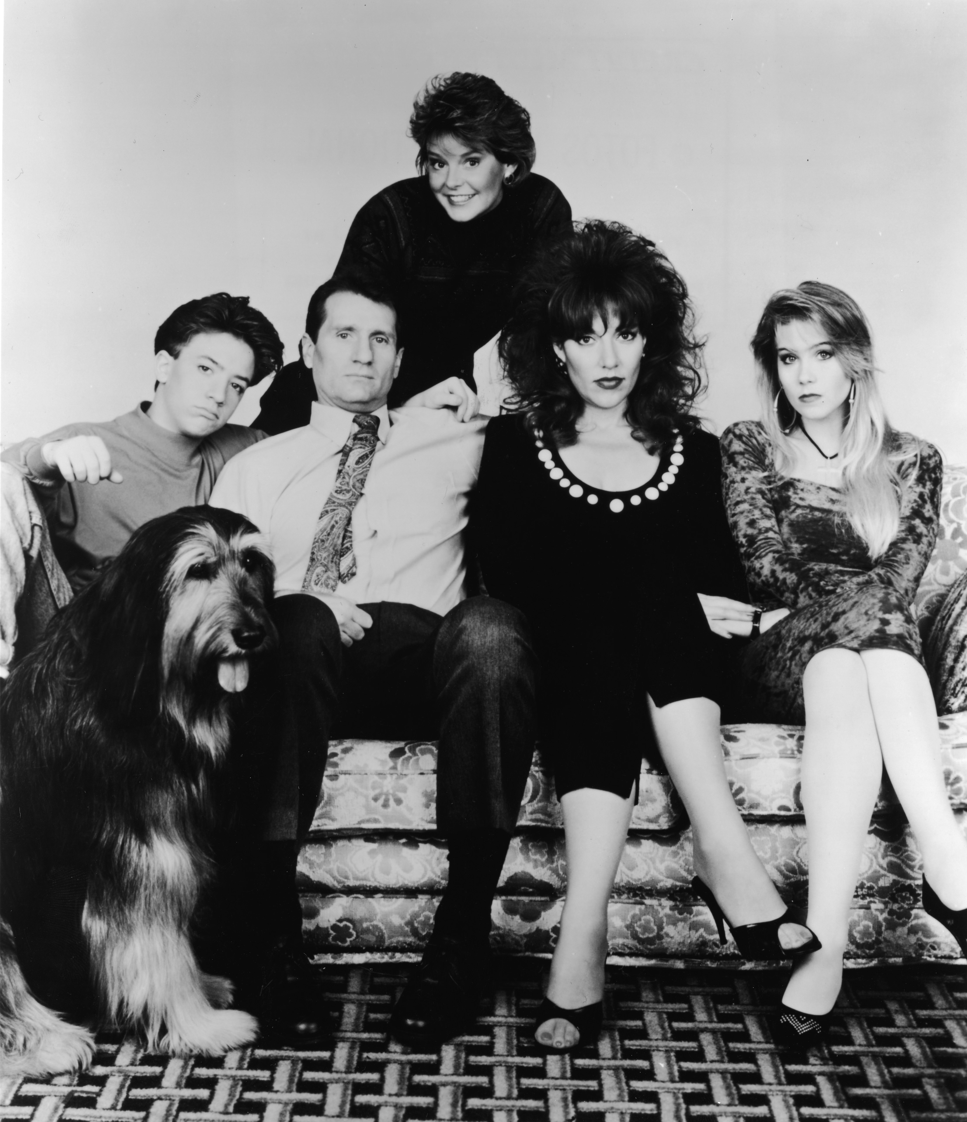 The cast of "Married... with Children"—including David Faustino, Ed O'Neill, Katey Sagal, Christina Applegate, and Amanda Bearse—posed alongside their long-haired family dog, Buck, for a classic black-and-white promotional portrait circa 1990. Squeezed onto the Bundy family's floral couch, O'Neill, Sagal, Faustino, and Applegate maintained deadpan, unenthusiastic expressions true to their cynical characters, while Bearse peered cheerfully over the sofa back. | Source: Getty Images