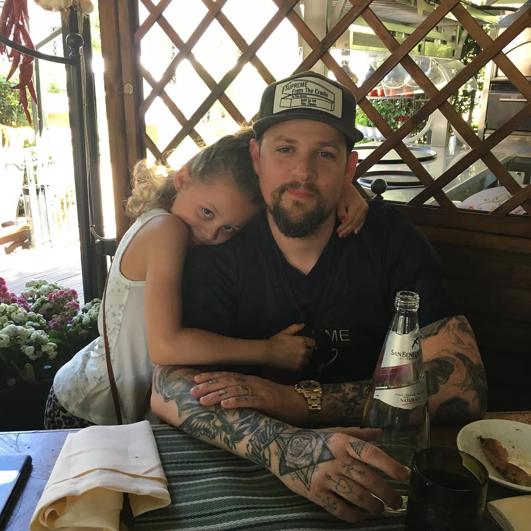 Joel Madden sits at a restaurant table while Harlow Winter Kate Madden leans into him with her arm around his shoulders. The candid image captures a warm father-daughter moment during Kate's childhood. | Source: Instagram/nicolerichie and joelmadden