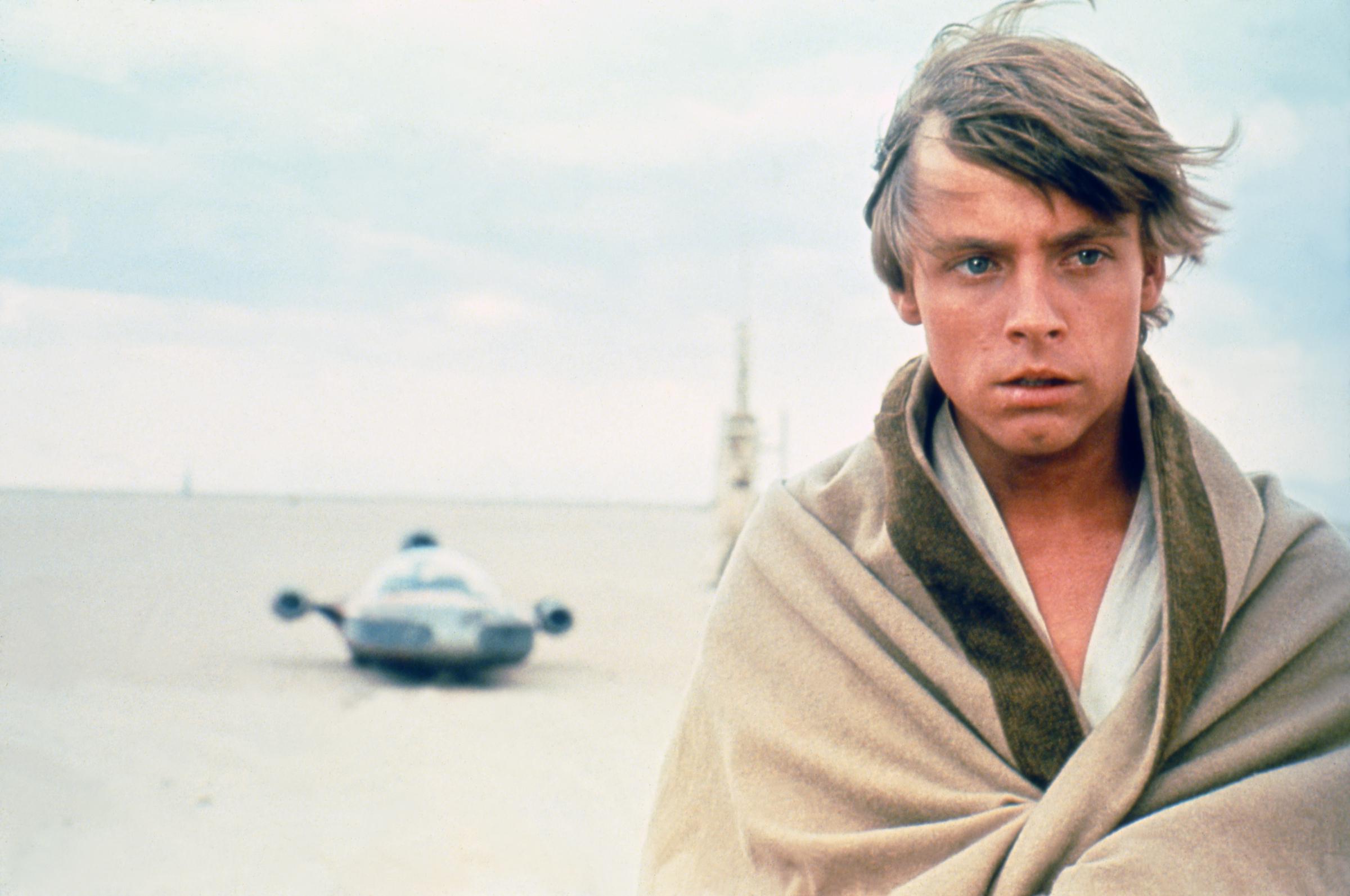 Mark Hamill stood against the stark desert landscape as Luke Skywalker during production of "Star Wars: A New Hope," with the character's landspeeder visible in the distance. Wrapped in light-colored clothing beneath the open sky, Hamill embodied the young hero before audiences knew his name. The production still provided another clear view of his appearance before the January 1977 accident that resulted in reconstructive treatment for his facial injuries.