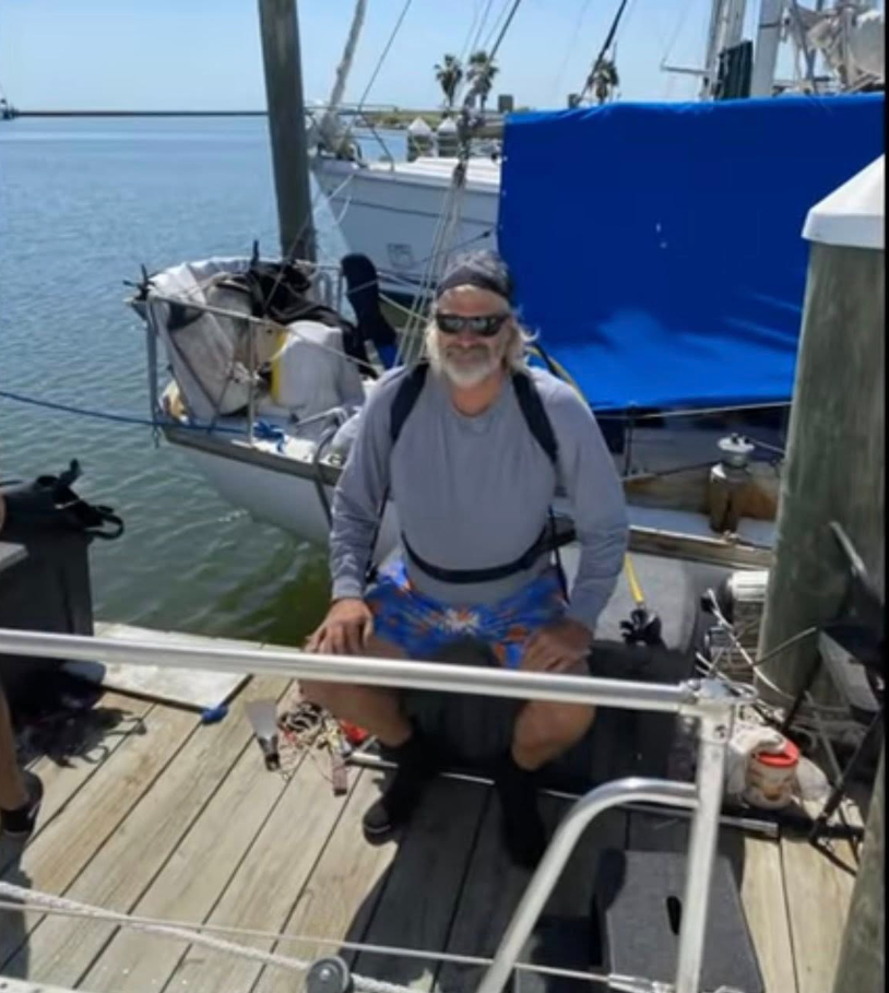 Brian Hooker is seen smiling on a dock beside a sailboat, wearing sunglasses, a cap, and casual boating gear. | Source: Facebook/PlunderStudios