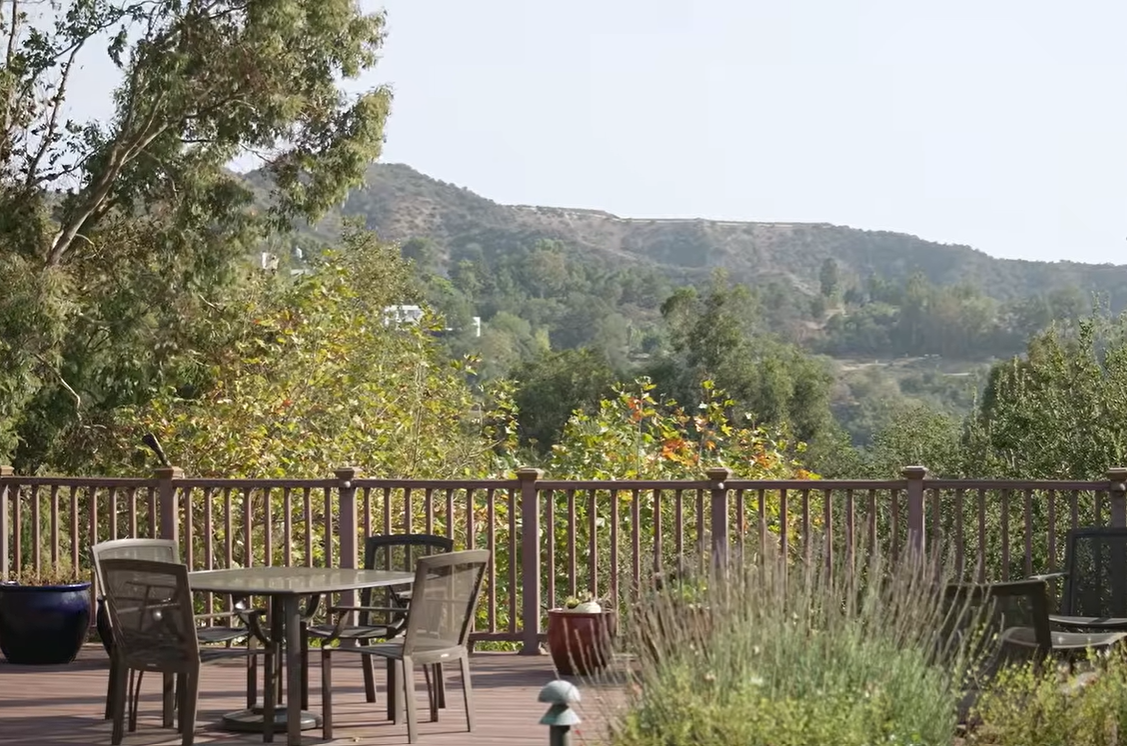 A wide view of the wooden deck at Barbara Eden's home reveals outdoor dining furniture, potted plants, and a railing overlooking lush trees and rolling hills beneath a clear sky. | Source: YouTube/People