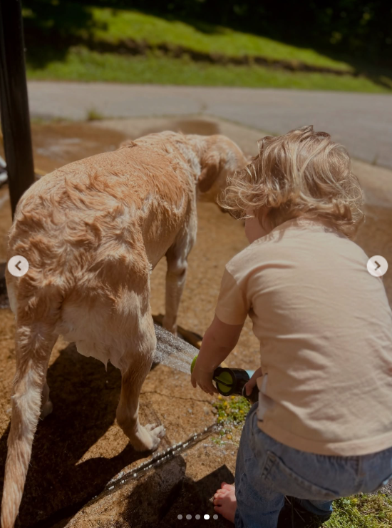 Kaliyah crouches beside a light-colored dog on a wet outdoor surface, holding a hose near the ground. The photograph adds another animal-care activity to the collection of moments from her childhood. | Source: Instagram/wynonnajudd