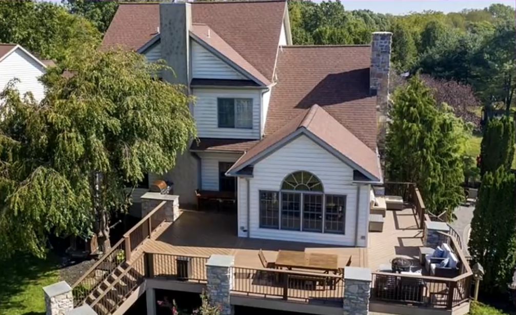 An elevated view shows the back of the Gosselin family home, with its multiple gables, brown shingle roof, stone chimneys, and expansive wooden deck surrounded by mature trees. The deck features outdoor seating, a dining table, and stairs leading down to the yard.