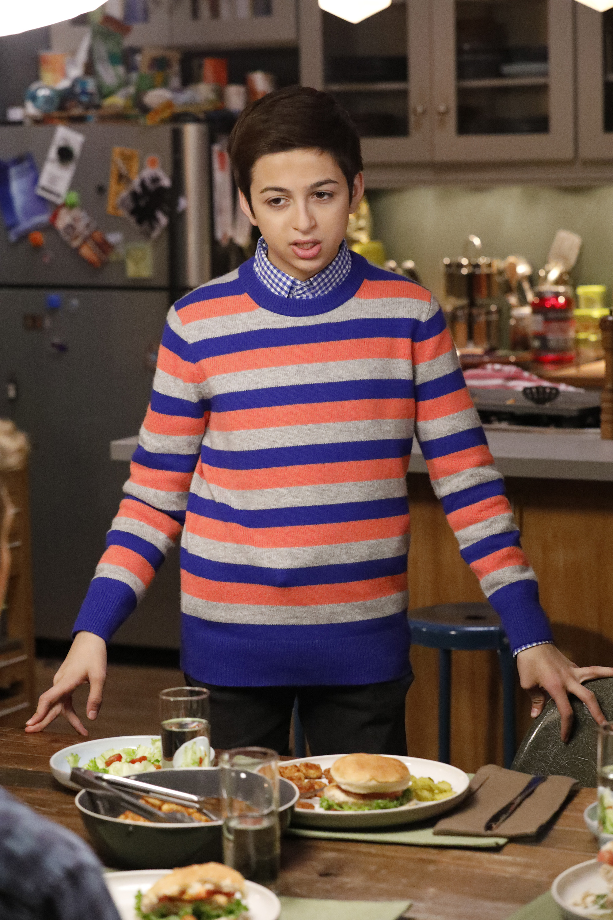 J.J. Totah portrays Michael in the Champions episode "Vincemas," wearing a colorful striped sweater over a blue checkered shirt while standing beside a dining table in a kitchen setting. | Source: Getty Images