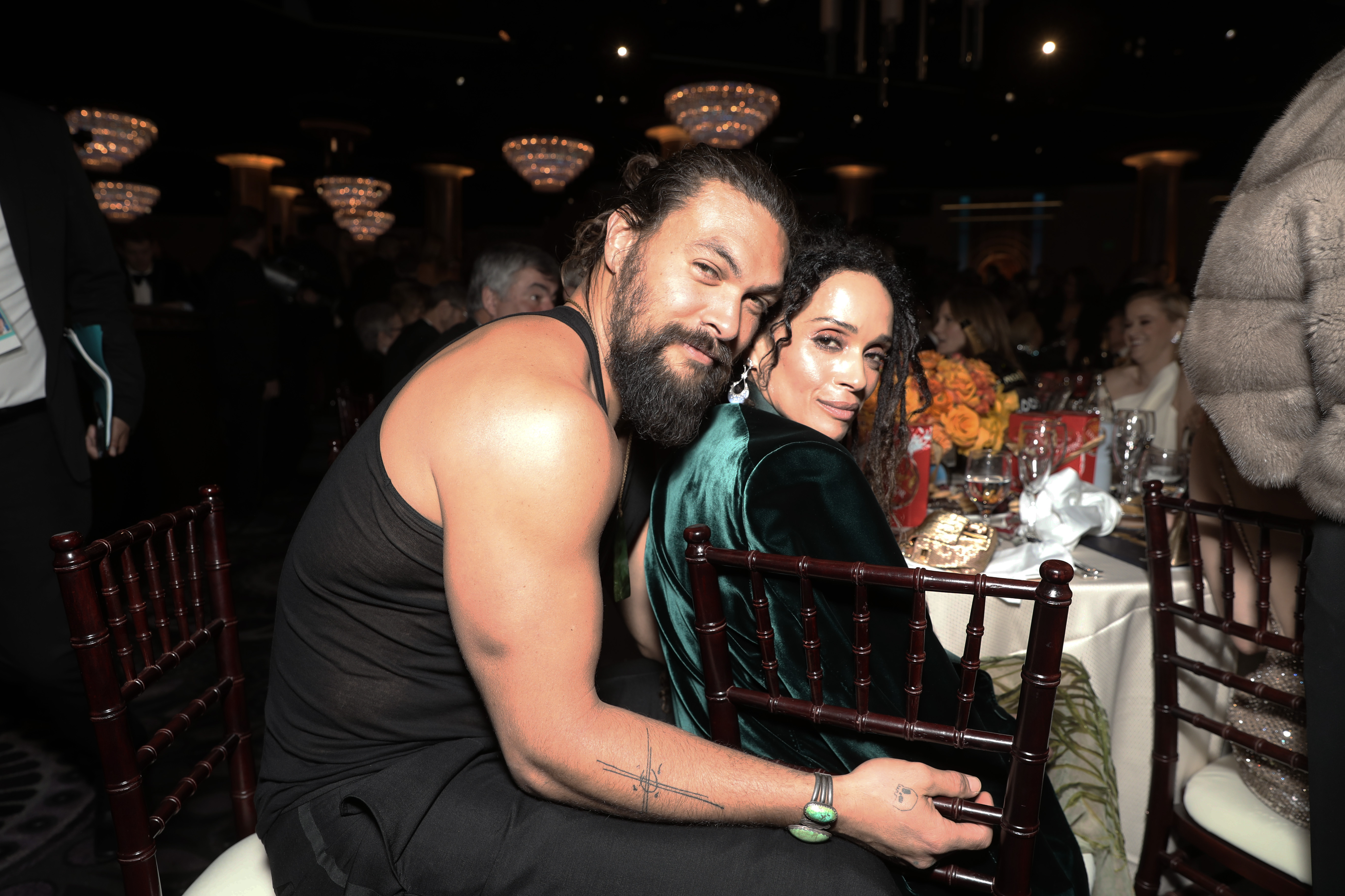 Jason Momoa and Lisa Bonet share an intimate moment seated together inside the ballroom at the 77th Annual Golden Globe Awards, held at The Beverly Hilton on January 5, 2020, in Beverly Hills, California. Momoa turns over his chair in a black ribbed tank top showcasing his arm tattoos and jade cuffs, resting an affectionate arm behind Bonet, who leans in while wearing his emerald-green velvet tuxedo jacket over her shoulder. | Source: Getty Images