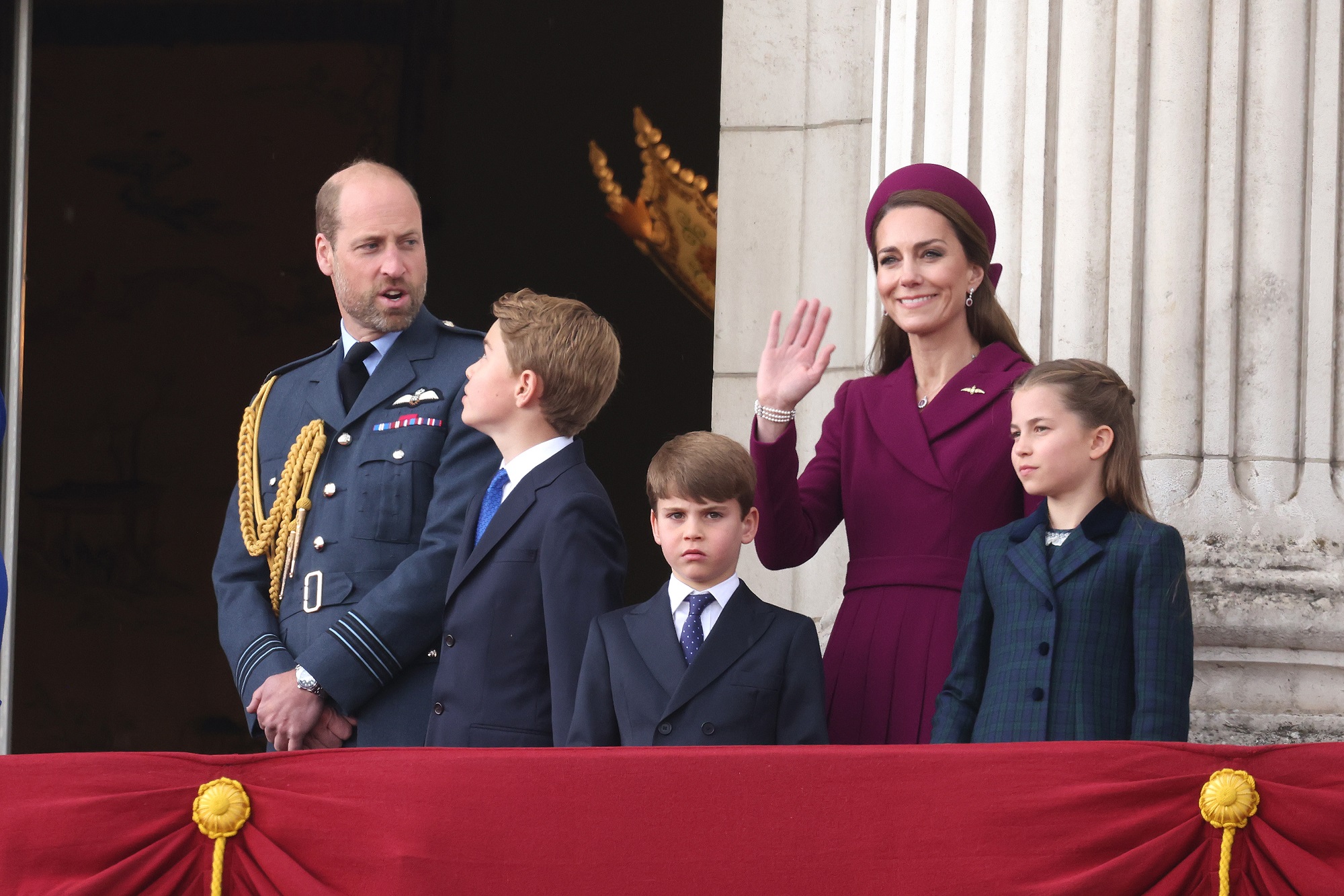 William, Catherine, and their children stand on the balcony. William appears to be speaking to George, while Catherine smiles and waves. Louis looks serious, and Charlotte gazes ahead with a composed expression.
