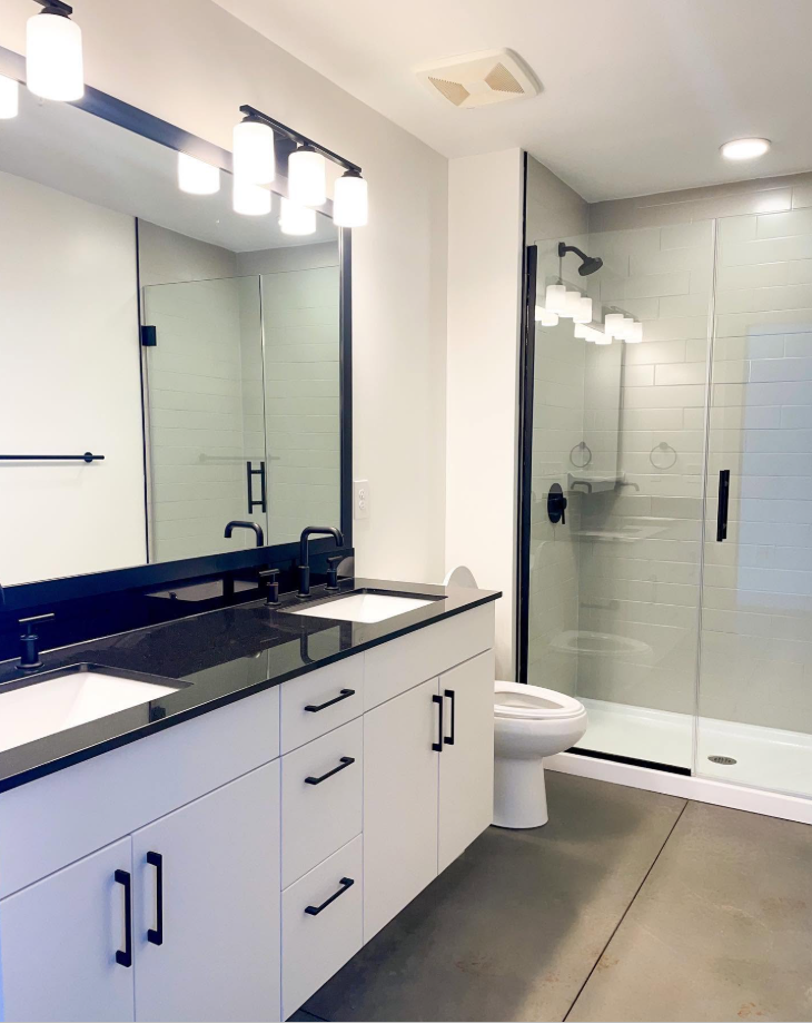 A bright bathroom pairs a generous double vanity with a glass-enclosed shower, bringing together white cabinetry, dark countertops and black fixtures. | Source: Facebook/Judson Mills Loft