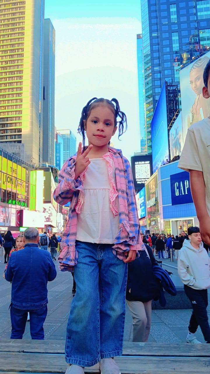 Eddimar Paola Hernández Chacón posed playfully, flashing a peace sign while standing outdoors in the bustling center of Times Square in New York City. Sporting pigtails, a white top layered under a pink-and-blue ruffled plaid button-down, and wide-leg denim jeans, she stood elevated before the iconic glowing billboards, towering skyscrapers, and busy pedestrian crowds. | Source: Facebook/Morena Esa
