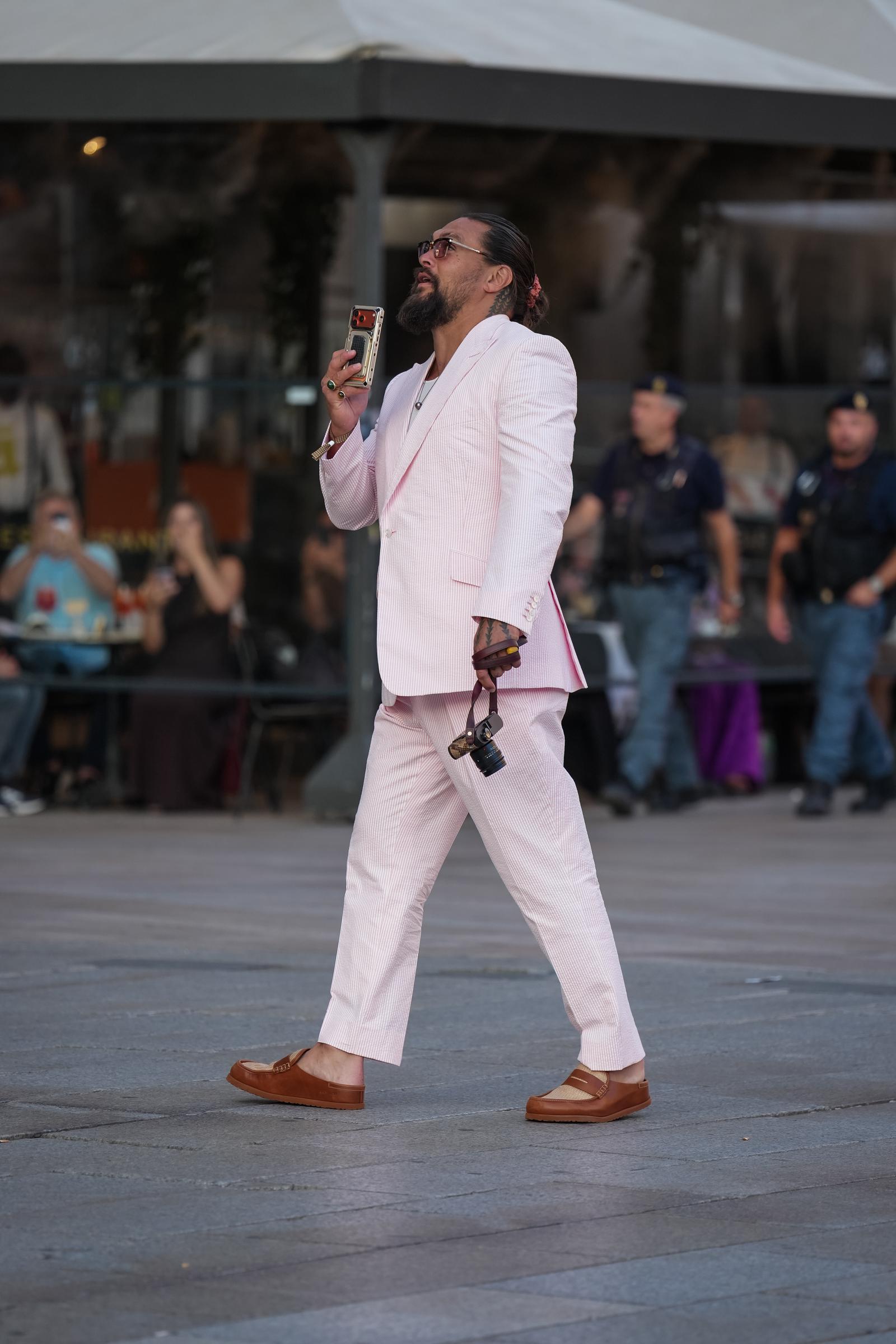 Momoa walks outside while holding up his phone, revealing the suit's relaxed proportions and brown slip-on shoes. | Source: Getty Images