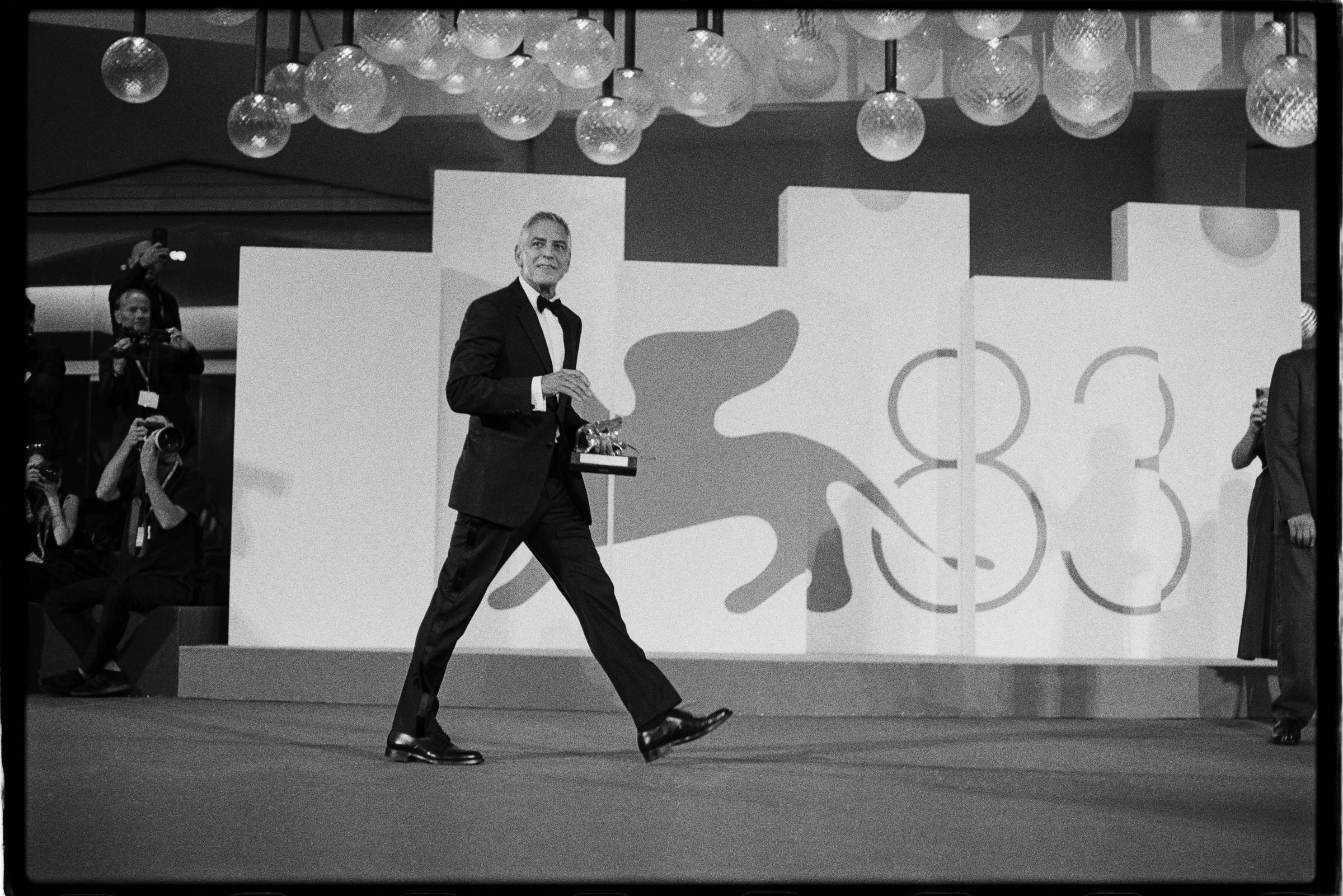 Captured in classic black and white on a vintage analogue camera, George Clooney proudly held his Golden Lion for Lifetime Achievement at the 83rd International Venice Film Festival on September 2, 2026, at Venice Lido. The acclaimed actor and filmmaker strode across the festival stage in a dapper tuxedo, bow tie, and patent leather dress shoes, smiling broadly as he carried the prestigious statuette past the event's backdrop. | Source: Getty Images