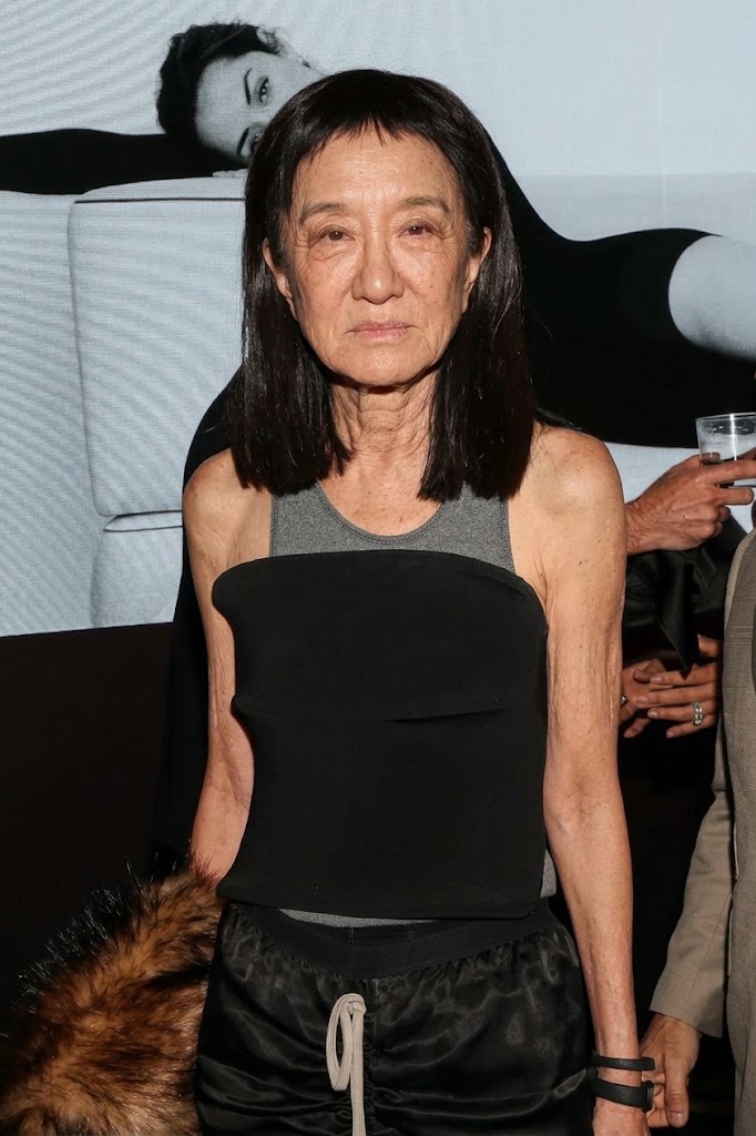 This AI-generated visualization depicts Vera Wang sporting blunt micro-bangs and straight dark hair, emphasizing realistic facial character lines and an unaugmented profile. Set against a monochrome interior backdrop, the simulated styling pairs casual streetwear influence with evening glamour, featuring a strapless black bodice layered over a heather-gray athletic tank and ruched drawstring separates. | Source: Gemini