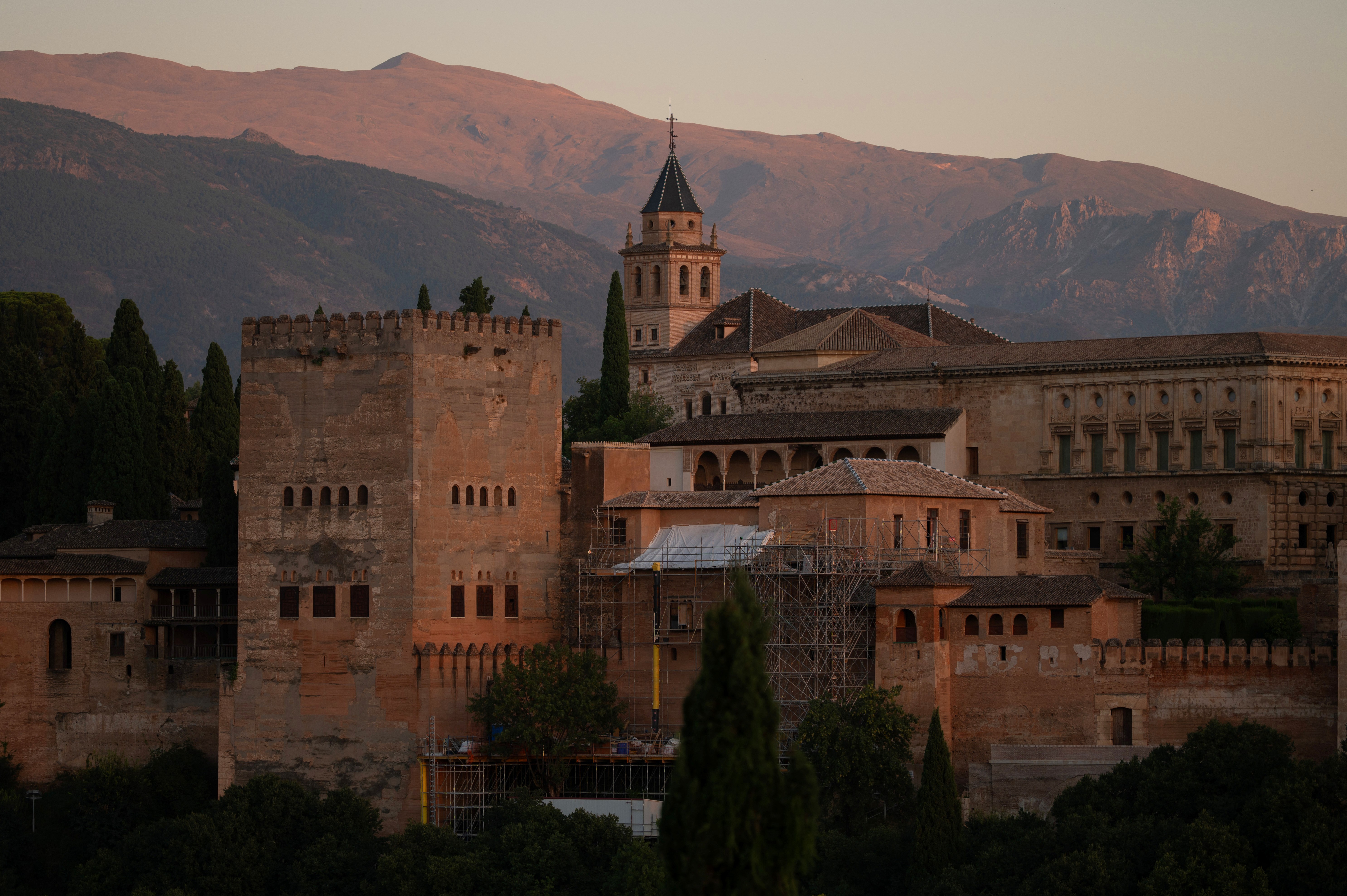 A scenic view captured the historic fortress and towers of the Alhambra from Saint Nicholas square following an earthquake in Granada on August 18, 2026. The ancient reddish stone architecture, church tower, and ongoing restoration scaffolding stood bathed in warm twilight against the towering backdrop of the Sierra Nevada mountains. | Source: Getty Images