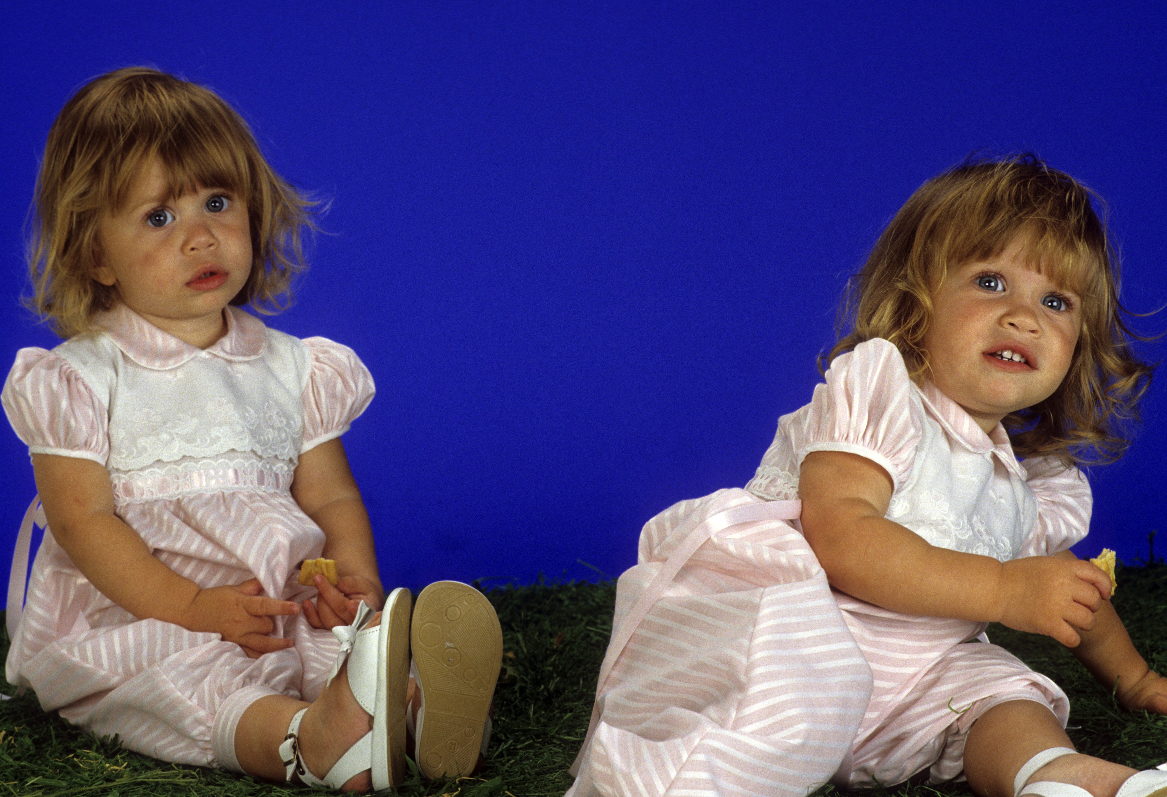 Round baby cheeks and wide, innocent eyes gave one-year-old Mary-Kate and Ashley Olsen a classic cherubic look during an April 11, 1988 gallery shoot for "Full House", complemented by matching pink-and-white striped rompers with puffed sleeves, Peter Pan collars, and tiny white buckle sandals.