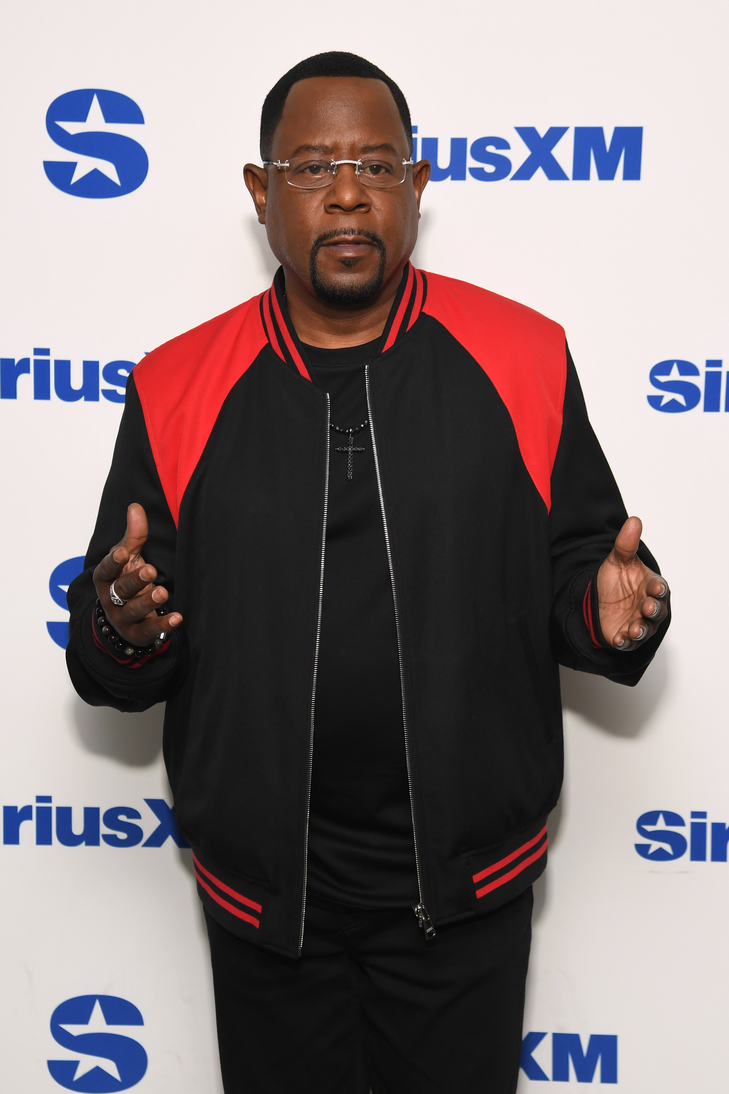 Still at SiriusXM Studios, Martin Lawrence stands in front of a branded backdrop in a black-and-red jacket. With both hands slightly raised, he looks focused and composed, striking a straightforward pose.