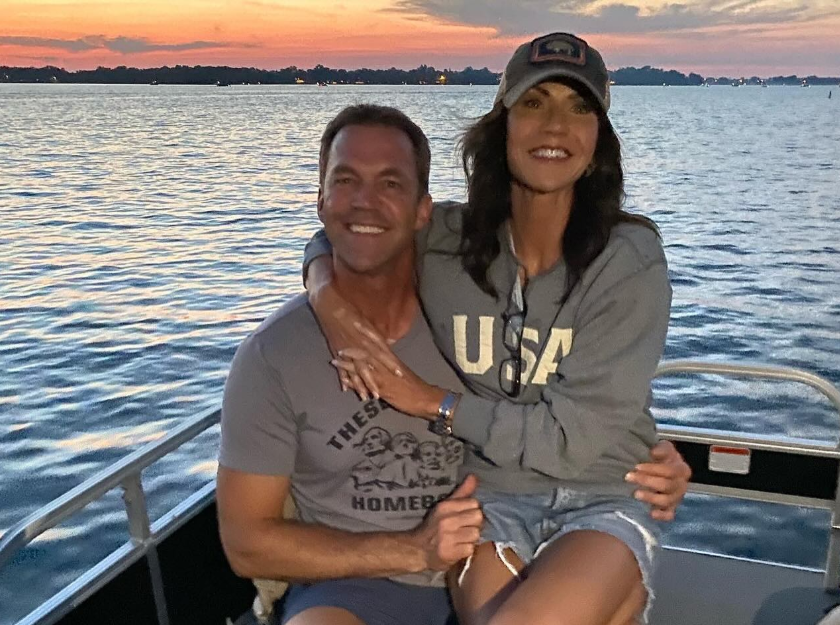 Kristi and Bryon Noem sit close on a boat at sunset, arms wrapped around each other as the water glows behind them—easy smiles, soft light, and a moment that feels both intimate and unguarded. | Source: Instagram/sdbryonnoem