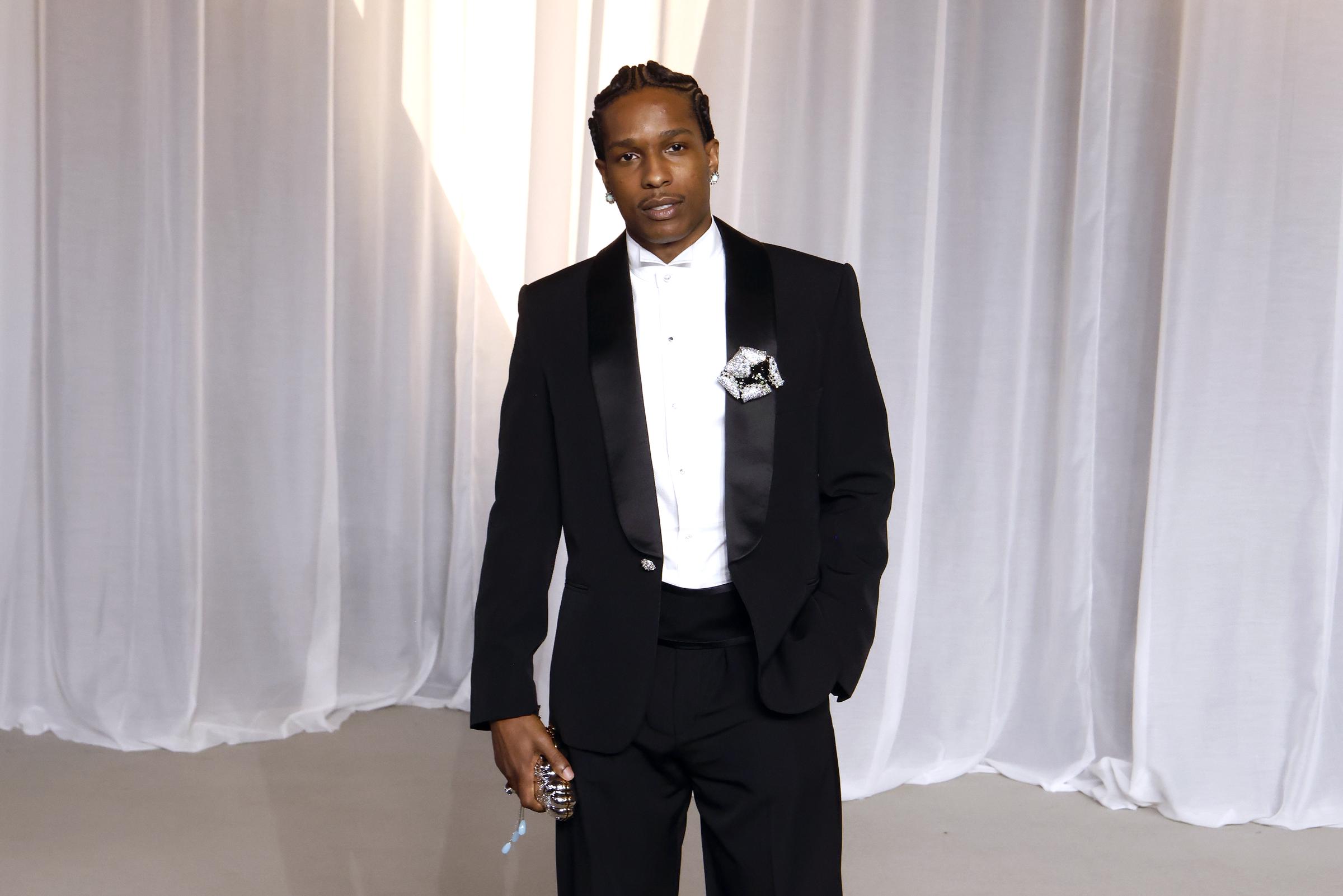 A$AP Rocky delivers modern black-tie sophistication in a sleek tuxedo tailored with wide satin shawl lapels and a high cummerbund waistband. Skipping a traditional bow tie for a minimalist wing-collared white dress shirt, the rapper elevates the classic ensemble with an ornate floral brooch pinned to his lapel, neat cornrows, and a sparkling novelty evening clutch.