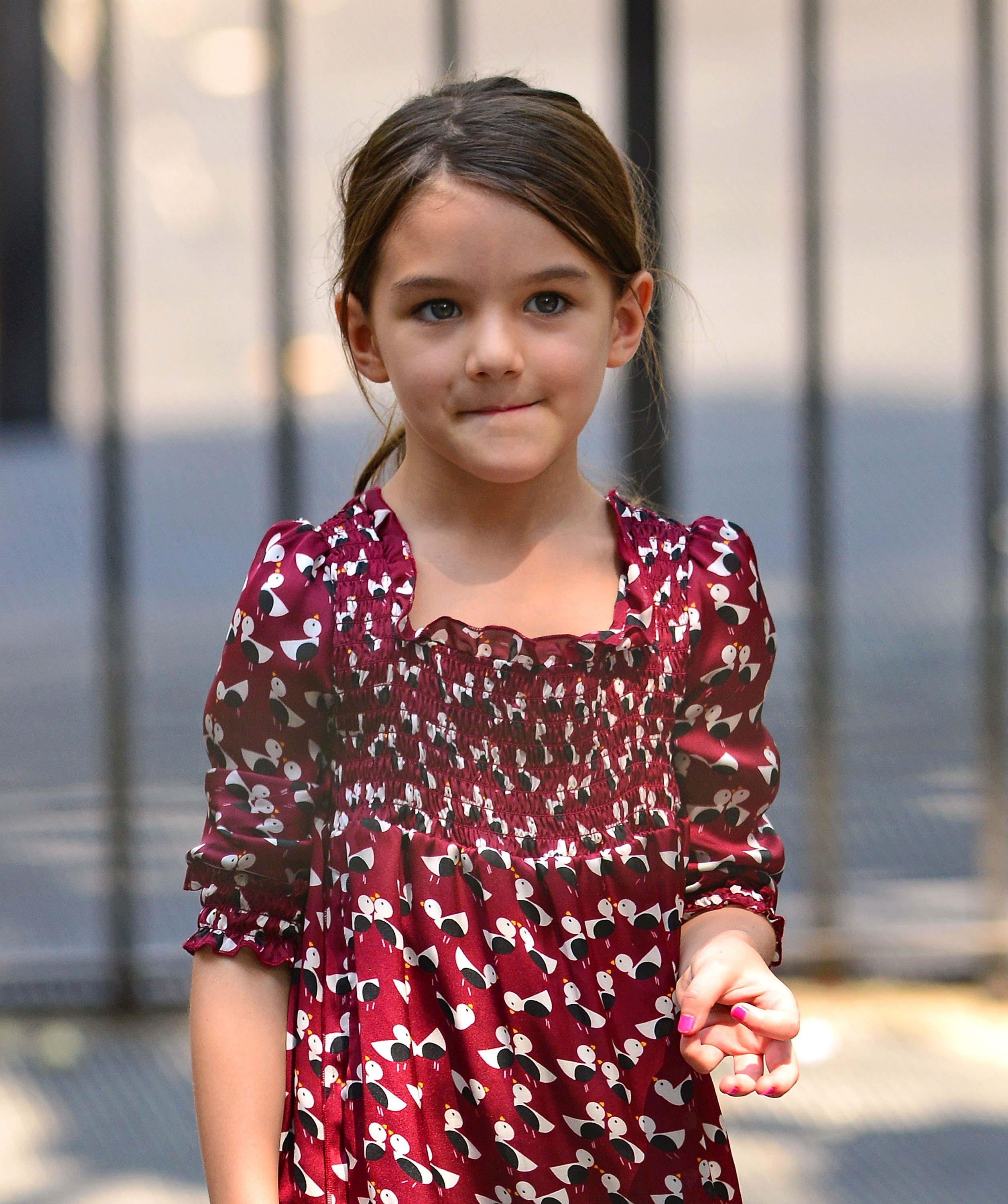 In this close-up street capture, young Suri Cruise wears a maroon dress that exudes her effortless charm. Her expression is calm and observant, offering a rare, unguarded look at the celebrity child beyond the headlines.