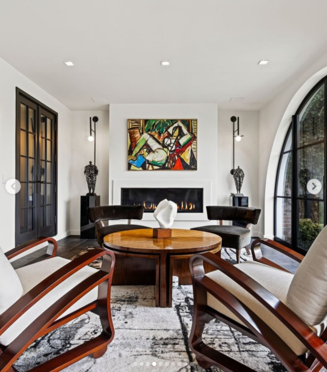 A brighter sitting area shows another side of the mansion, pairing white walls and an arched black-framed window with contemporary artwork and sculptural furniture. A sleek fireplace anchors the room, creating a sharp contrast with the estate's more traditional spaces. | Source: Instagram/rogerperrygroup
