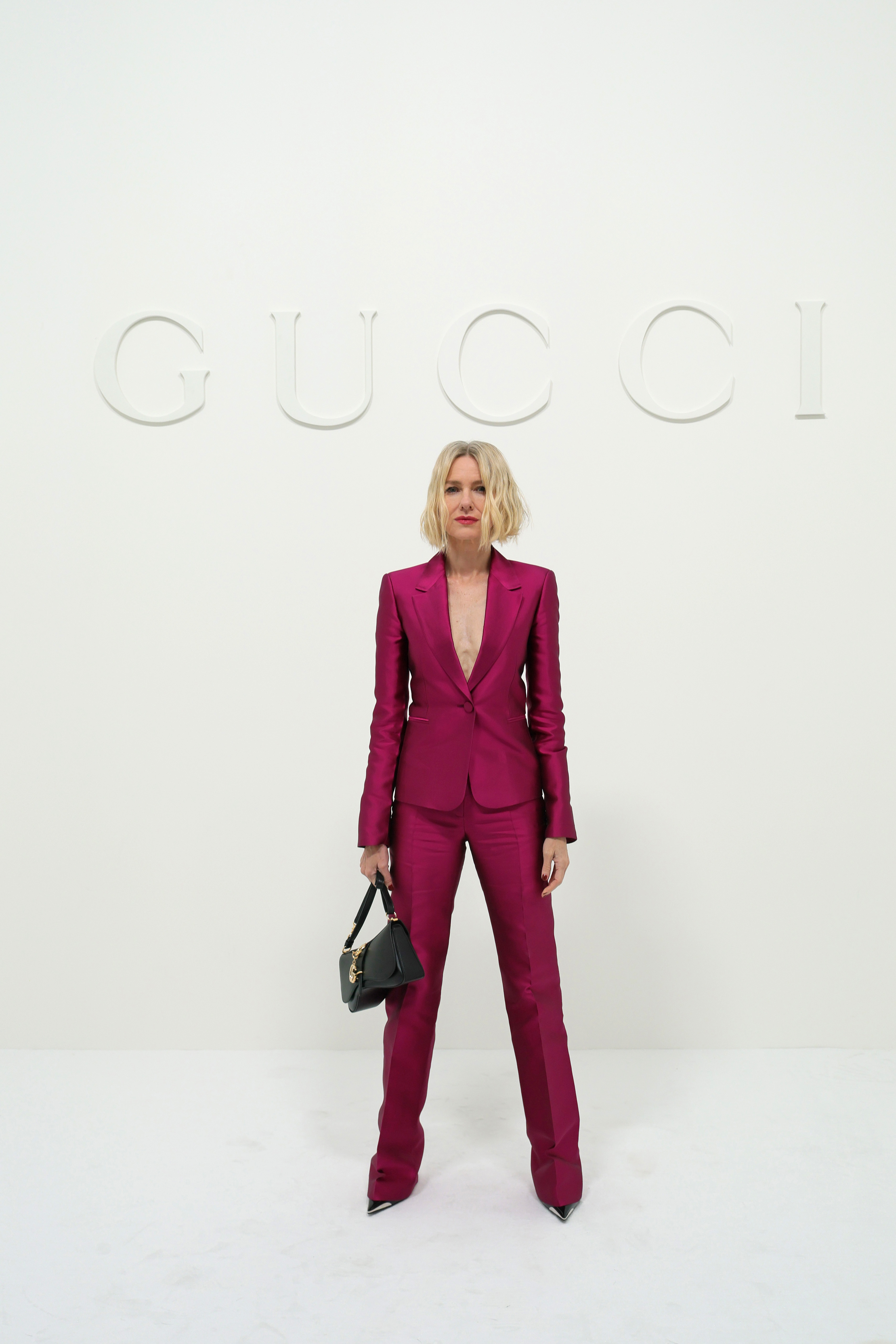Naomi Watts commanded the minimalist photo wall beneath a subtle white Gucci logo at Allianz MiCo on September 25, 2026, during Milan Fashion Week Womenswear Spring/Summer 2027 in Milan, Italy. Striking a confident pose with one hand carrying a structured black purse, she highlighted the sharp tailoring and rich fuchsia tone of her satin suit alongside textured, chin-length blonde locks. | Source: Getty Images
