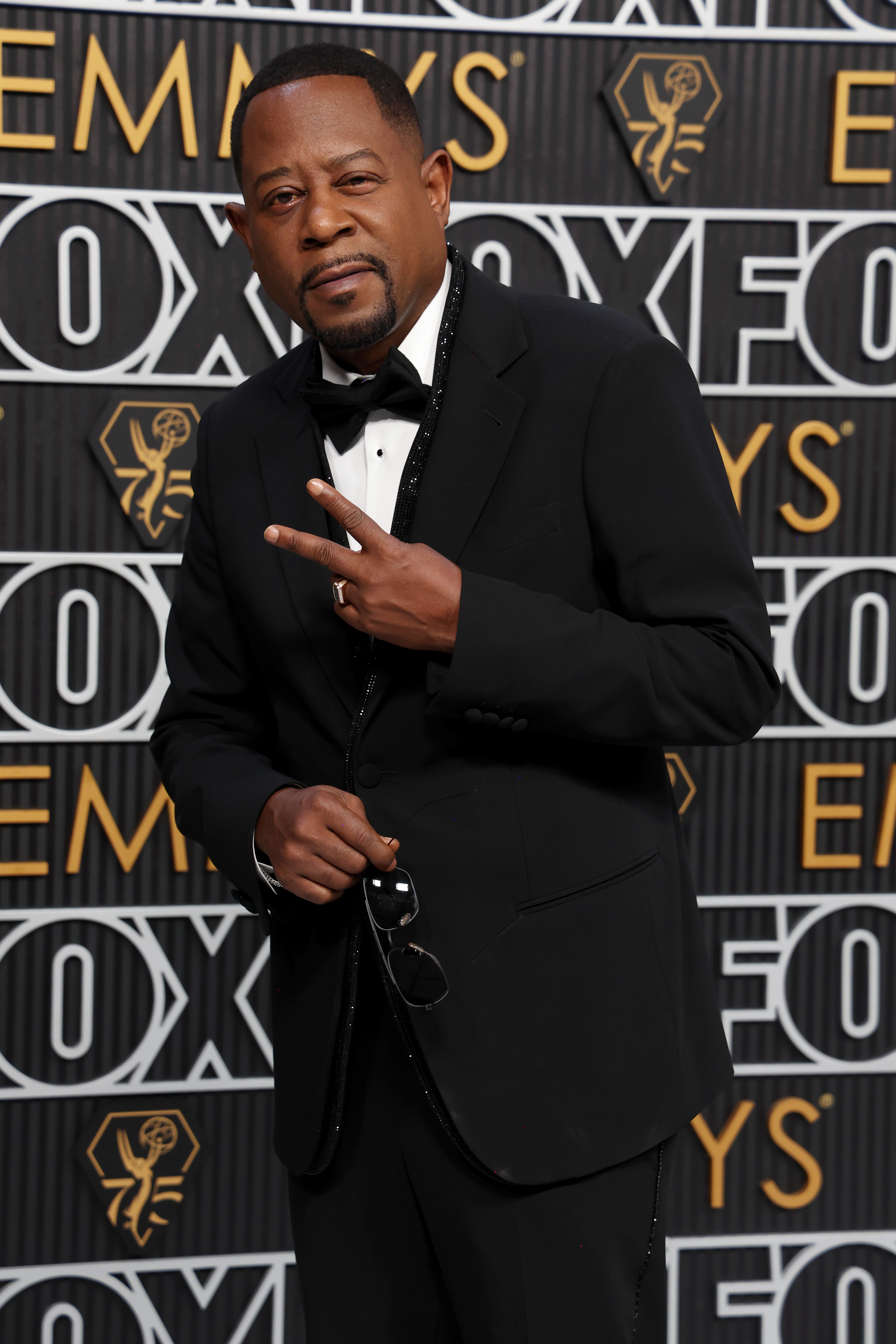 At the 75th Primetime Emmy Awards, Martin Lawrence wears a classic black tuxedo with a bow tie. He poses confidently, flashing a peace sign while holding his sunglasses, exuding a cool, celebratory energy against the branded backdrop.