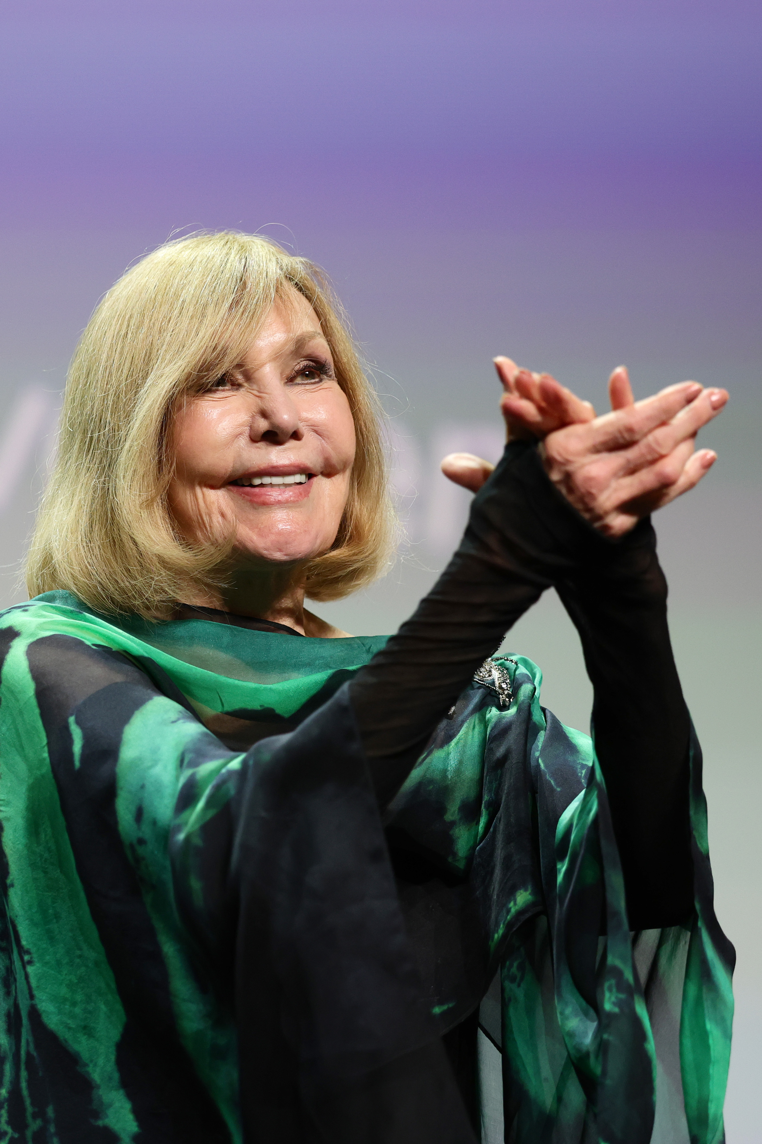 Kim Novak smiles and applauds during the Golden Lion for Lifetime Achievement ceremony at the 82nd Venice International Film Festival on September 1, 2025. She wears a flowing teal-and-green wrap over a black outfit as she acknowledges the audience. | Source: Getty Images