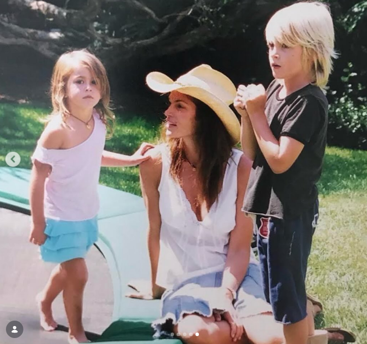 Cindy Crawford donned a wide-brimmed straw cowboy hat and a casual white top as she relaxed outdoors on the grass with her young children, Kaia and Presley, in a nostalgic family snapshot shared on February 20, 2024. | Source: Instagram/presleygerber