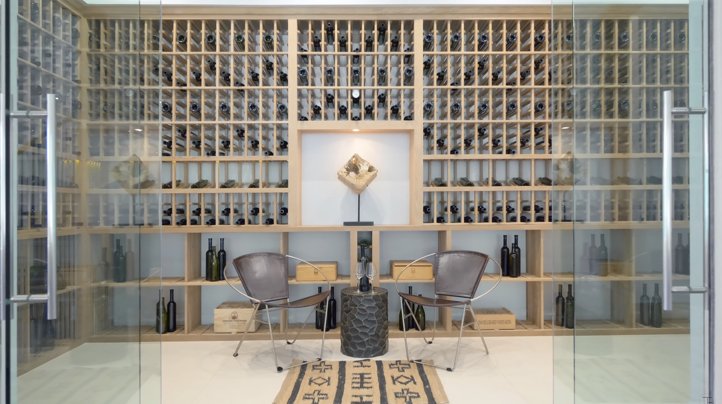 A glass enclosure keeps the extensive bottle storage visible from outside the room. Two chairs and a small table create a seating area against a wall of wood wine racks. | Source: Youtube/Tomer.Fridman