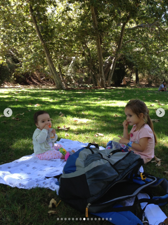 Mabel and baby Fiona share a quiet picnic in a wooded park, with Fiona sitting on a blanket and holding a snack while Mabel, wearing a pink T-shirt, sits nearby. A large blue-and-black stroller or baby carrier fills the foreground, capturing a candid moment from the sisters’ childhood. | Source: Instagram/mabellowe_