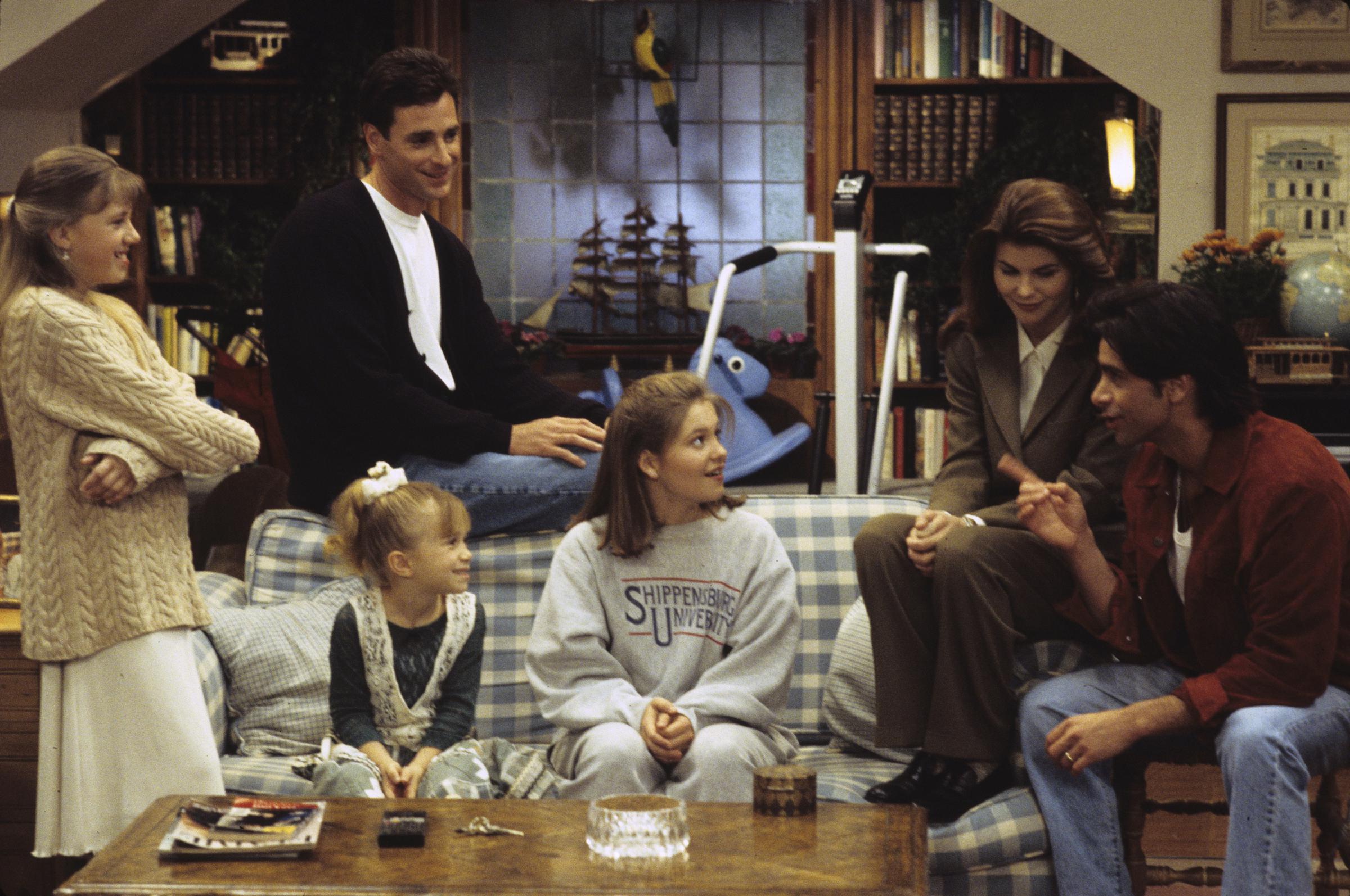 Jodie Sweetin, Candace Cameron Bure, Lori Loughlin, and John Stamos huddle in the living room with Bob Saget and Mary-Kate or Ashley Olsen in the Season 7 episode "Love on the Rocks," which aired on March 1, 1994. Cameron Bure lounges in comfortable casual fleece while Sweetin smiles from the side, listening as Stamos and a polished Loughlin help the family conspire against Joey's pranks. The scene captures the effortless camaraderie and playful warmth that kept "Full House" fans hooked through seven seasons of family hijinks.