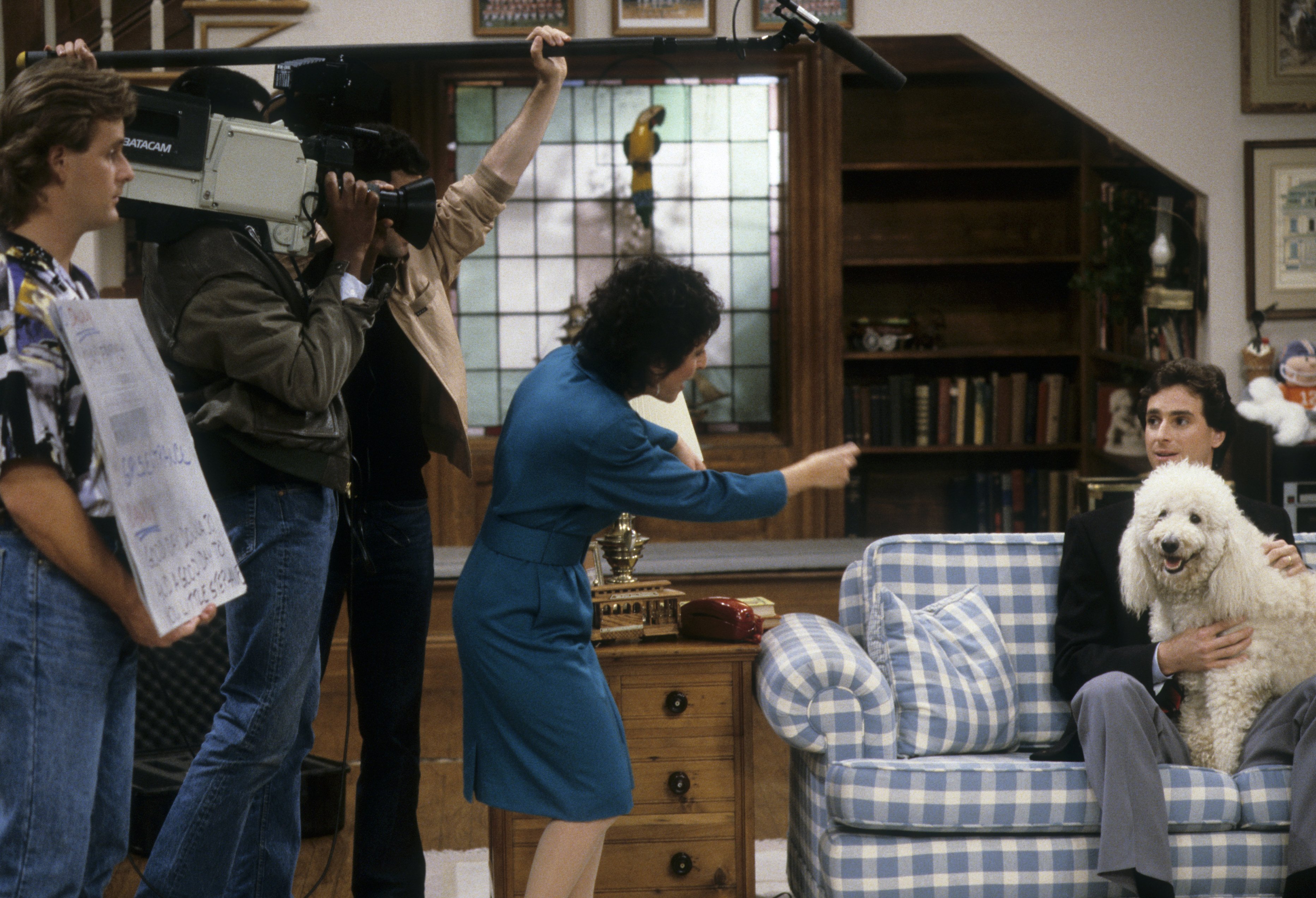 Bob Saget sits on the living room sofa with a white poodle as Belita Moreno directs him and Dave Coulier looks on during the Season 1 episode "Our Very First Promo," which aired on December 18, 1987. Capturing the meta humor of Danny Tanner's early television broadcast career, the scene highlights the foundational charm of "Full House" during its debut year, establishing the warm and chaotic household dynamics that would captivate audiences for decades to come.