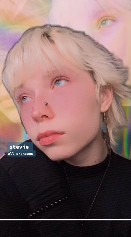 In a close-up shot, Stevie has platinum blonde hair and a nose piercing. She wears dark clothing with a soft, abstract background. The label updates to show she uses all pronouns | Source: TikTok/st0nedc0w