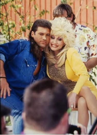 A candid, color photograph shows a younger Billy Ray Cyrus sporting his famous mullet in a denim shirt, leaning in close with Dolly Parton, who looks bright in a yellow outfit.