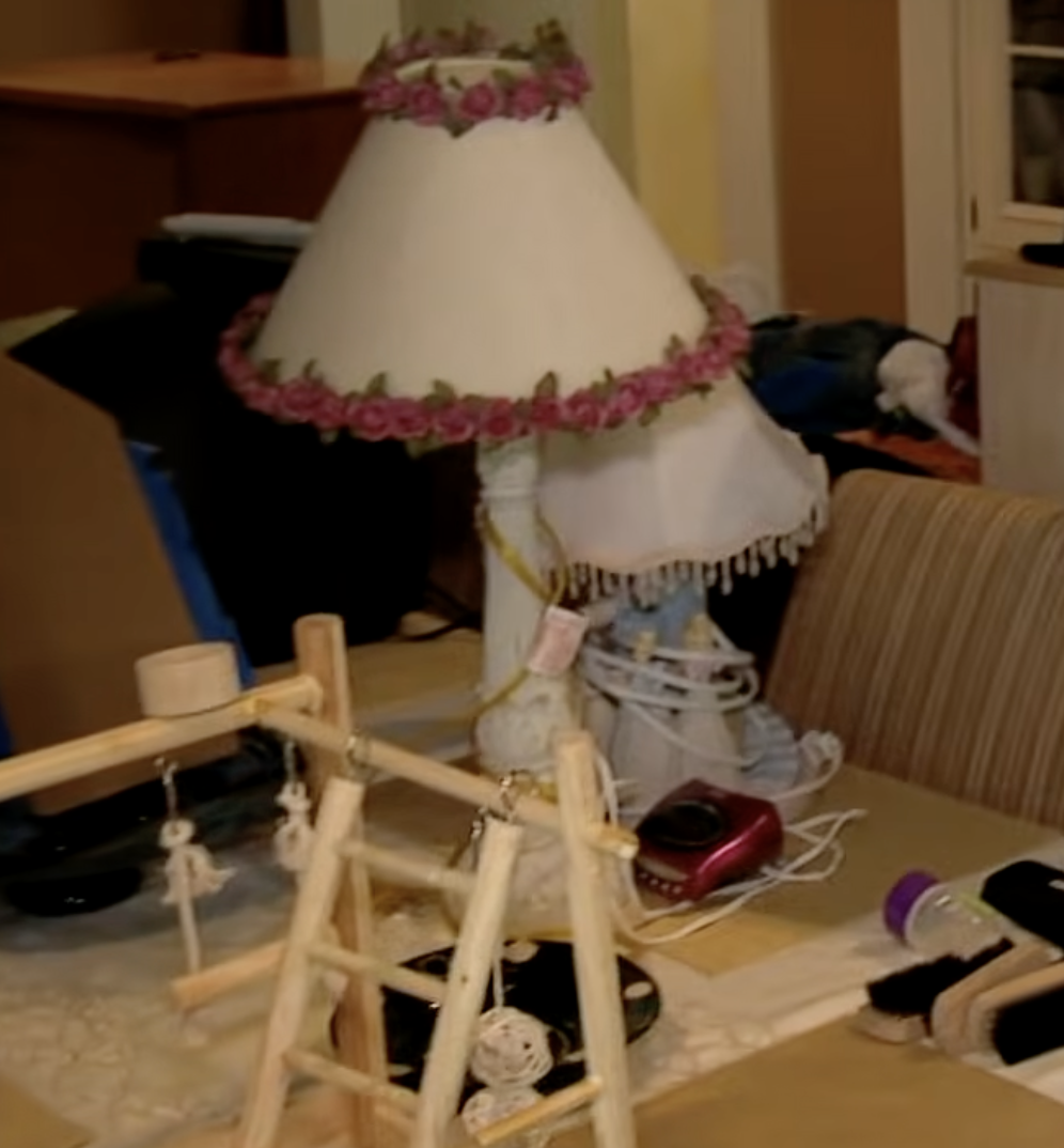 A cluttered tabletop in the Gosselin home holds a decorative lamp with pink floral trim, tangled electrical cords, small household items, and a wooden frame or stand during Kate Plus 8: Construction Prep. Boxes and other belongings fill the surrounding area as the family prepares for changes to the children's bedrooms. | Source: YouTube/TLC