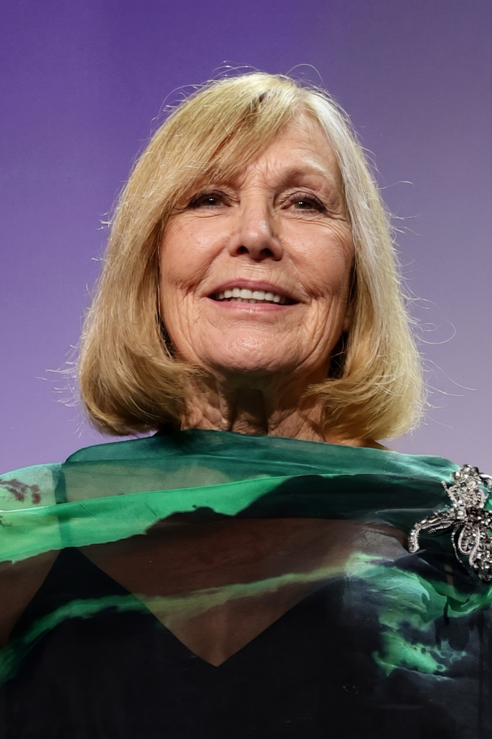 Kim Novak smiles warmly in a green-and-black ensemble featuring sheer fabric and a jeweled brooch on the shoulder, set against a soft purple background. | Source: Gemini