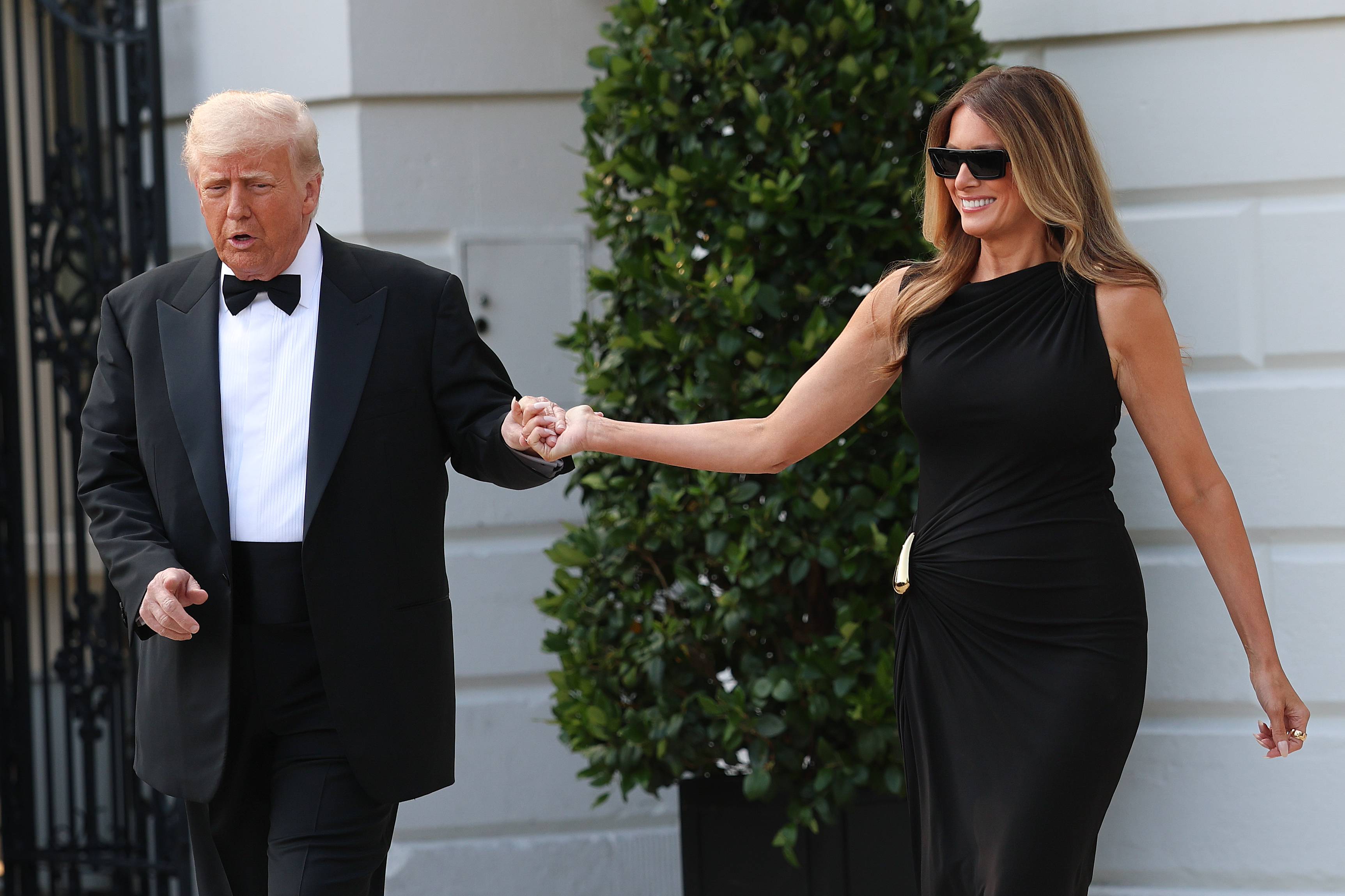 While the president maintains a serious expression, the first lady smiles brightly behind dark sunglasses as the couple exits the White House hand in hand on June 11, 2025.