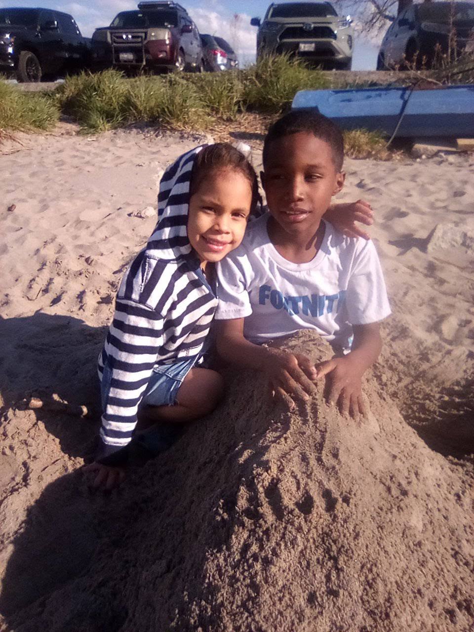 Eddimar Paola Hernández Chacón leaned affectionately against her brother while playing in a large mound of sand during a sunny beach trip. Smiling brightly with her hood up, Eddimar wore a navy-and-white striped zip-up hoodie over denim shorts, while her brother wrapped an arm around her shoulder in a white "Fortnite" graphic t-shirt. | Source: Facebook/Morena Esa