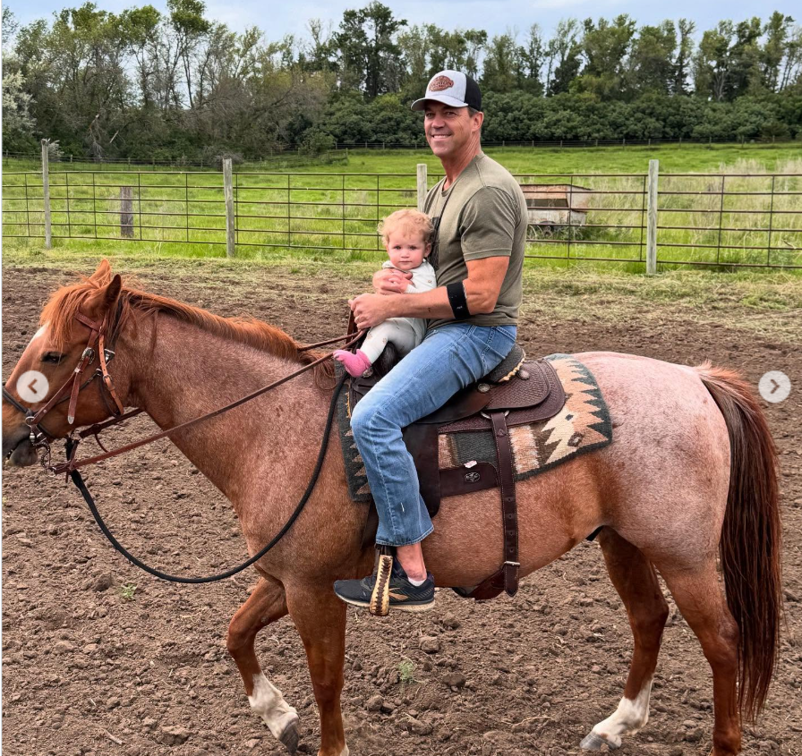 Bryon Noem rides a horse while holding a young child, both facing the camera in a rural setting with fencing and greenery in the background. | Source: Instagram/kristinoem