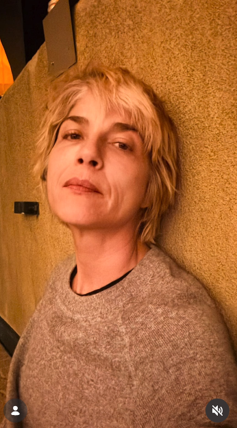 Selma Blair looks upward in a relaxed low-angle portrait, leaning her head against a textured earthen wall on October 11, 2026. Warm indoor ambient lighting casts a soft tone across her layered blonde crop and heathered grey crewneck knit. | Source: Instagram/selmablair