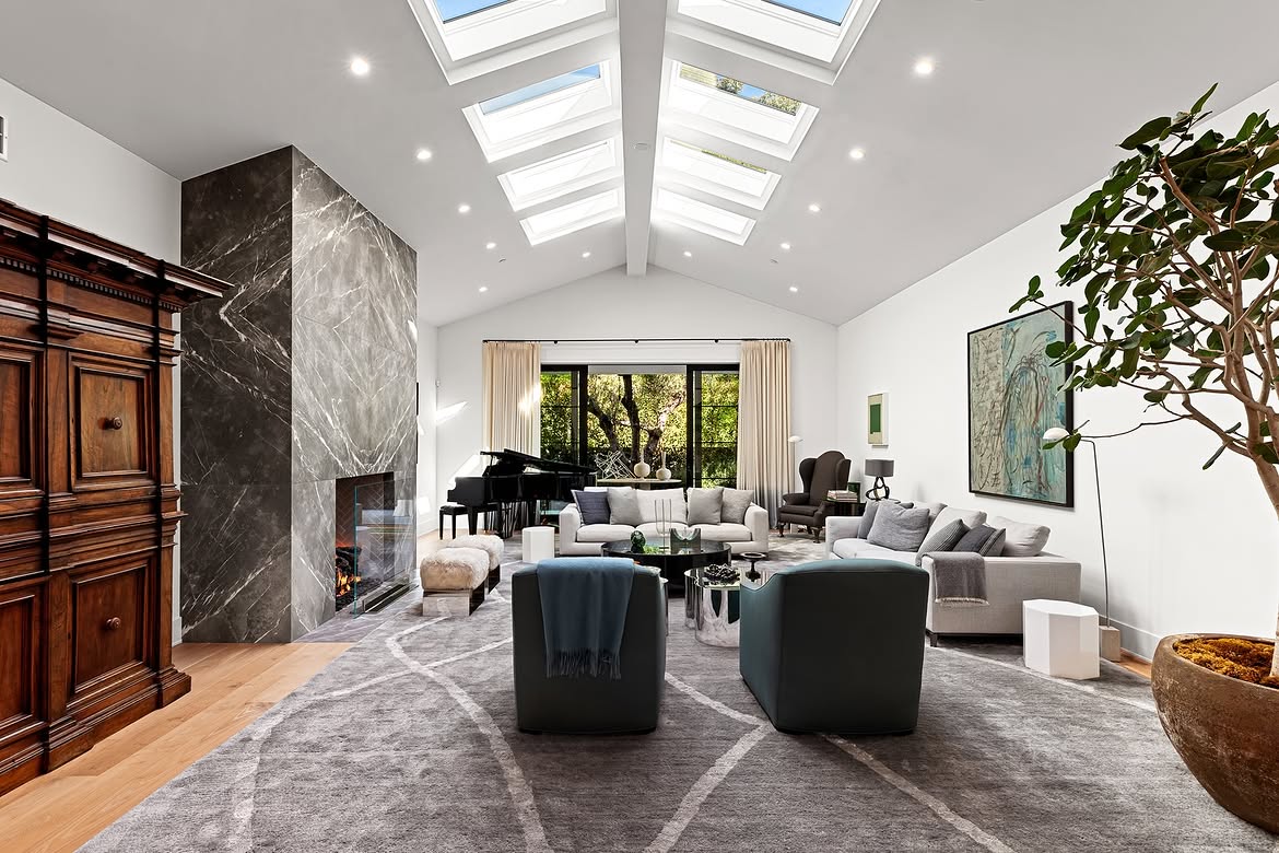 A second view of the formal living room highlights the long row of skylights above the vaulted ceiling. The dark stone fireplace, grand piano, and mature indoor tree add contrast to the room's otherwise pale backdrop. | Source: Instagram/tomer.fridman