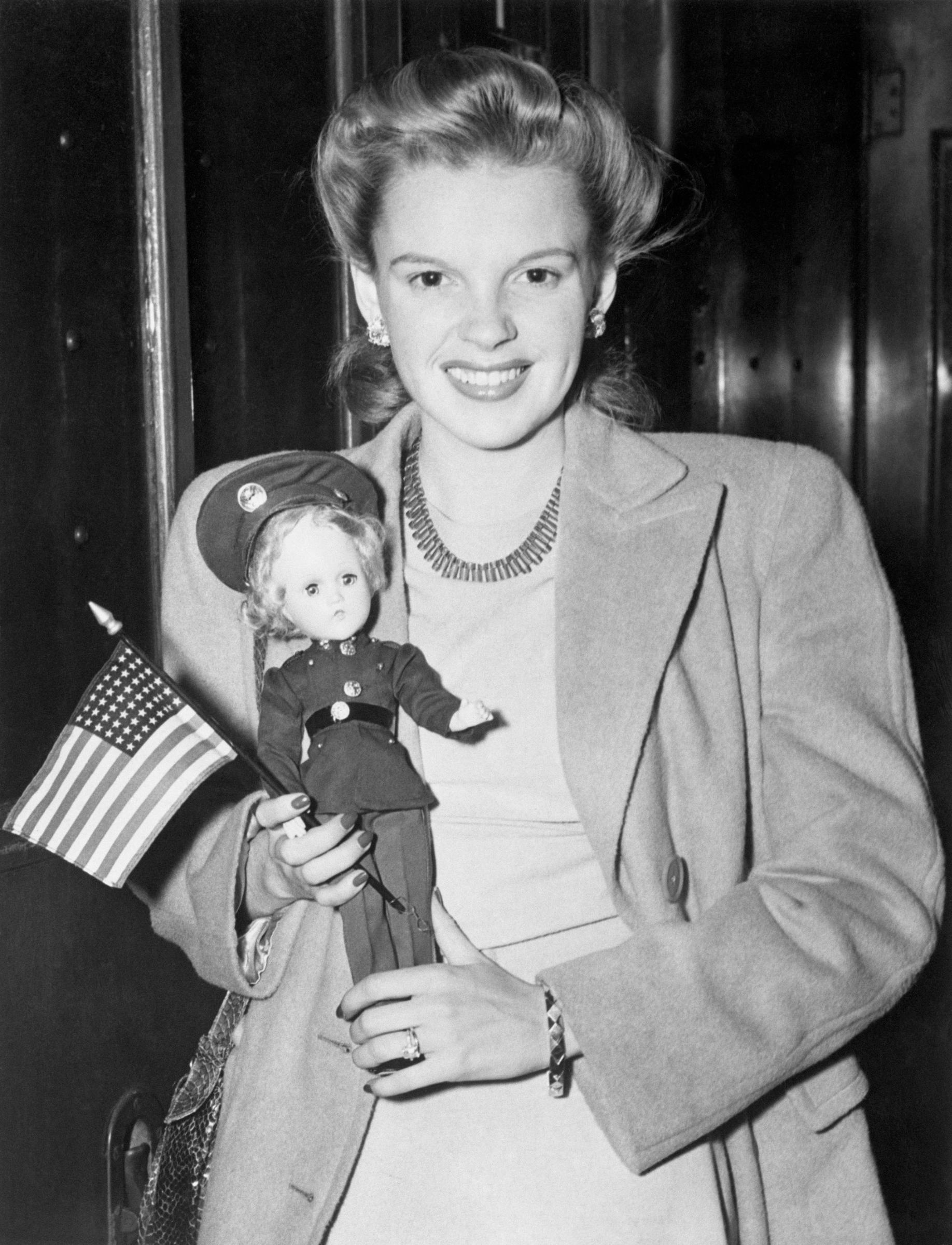 Judy Garland arrives in New York, holding a flag and doll while posing for photographers at a train station | Source: Getty Images
